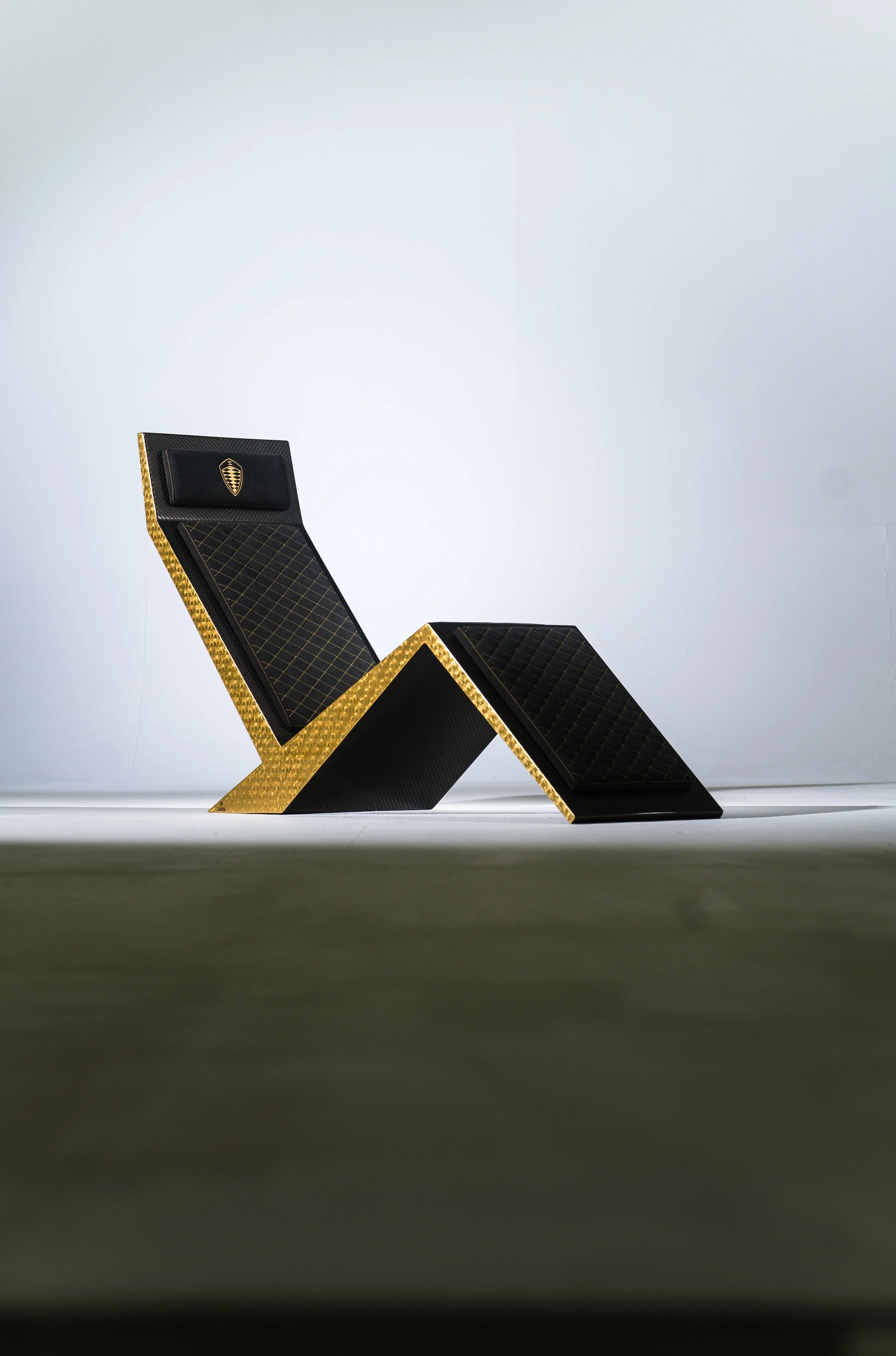 A modern black and gold lounge chair with a matching footrest, positioned on a plain floor against a white background.