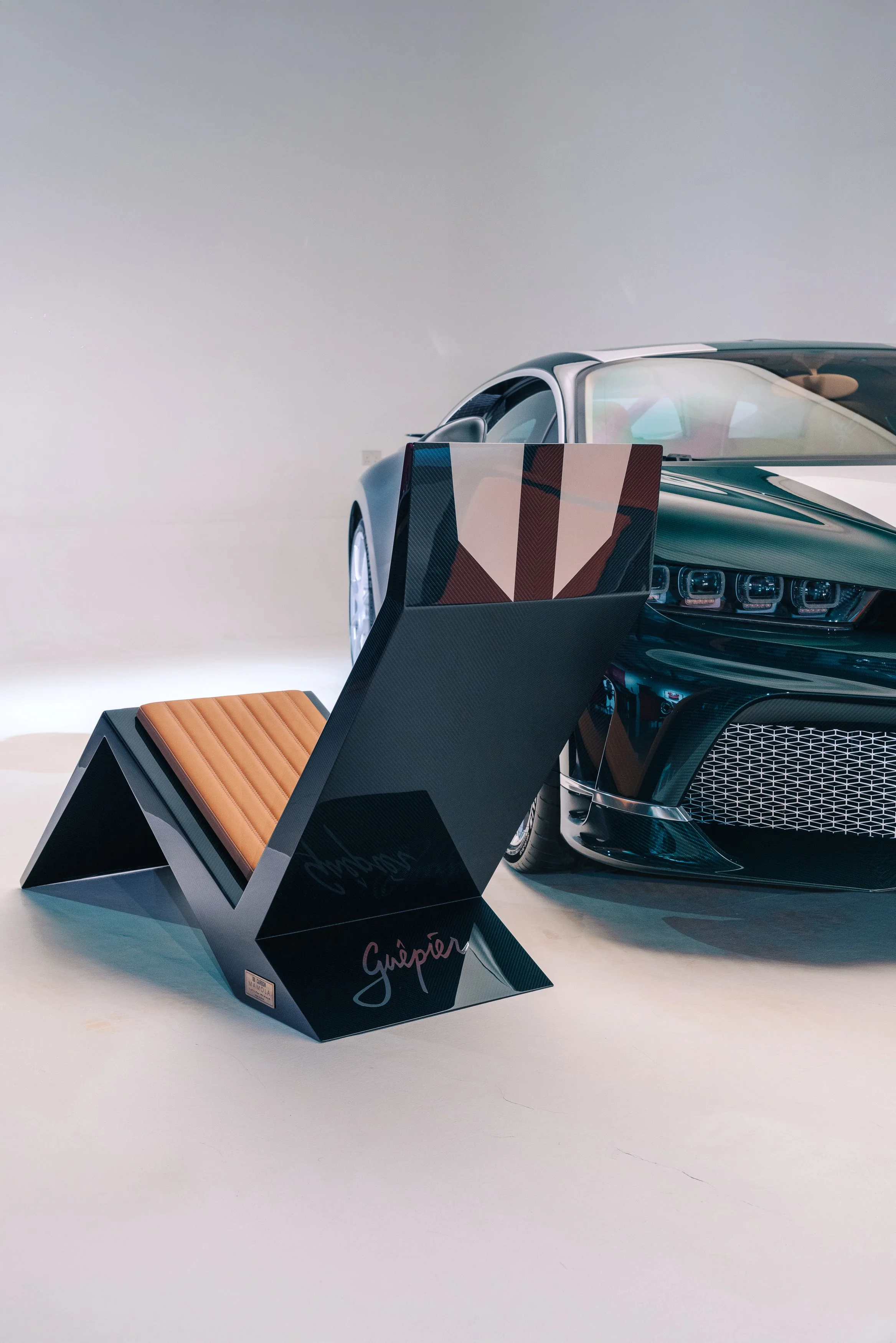 A black sports car with a sleek design and a distinctive front grille, partially visible, alongside a modern, artistic lounge chair with a copper-colored cushion, set against a plain white background.