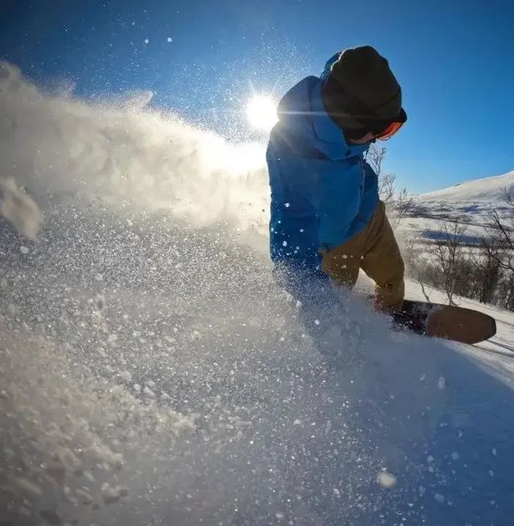 En snowboardåkare gör en kraftig sväng i lös pudersnö med solen i bakgrunden.