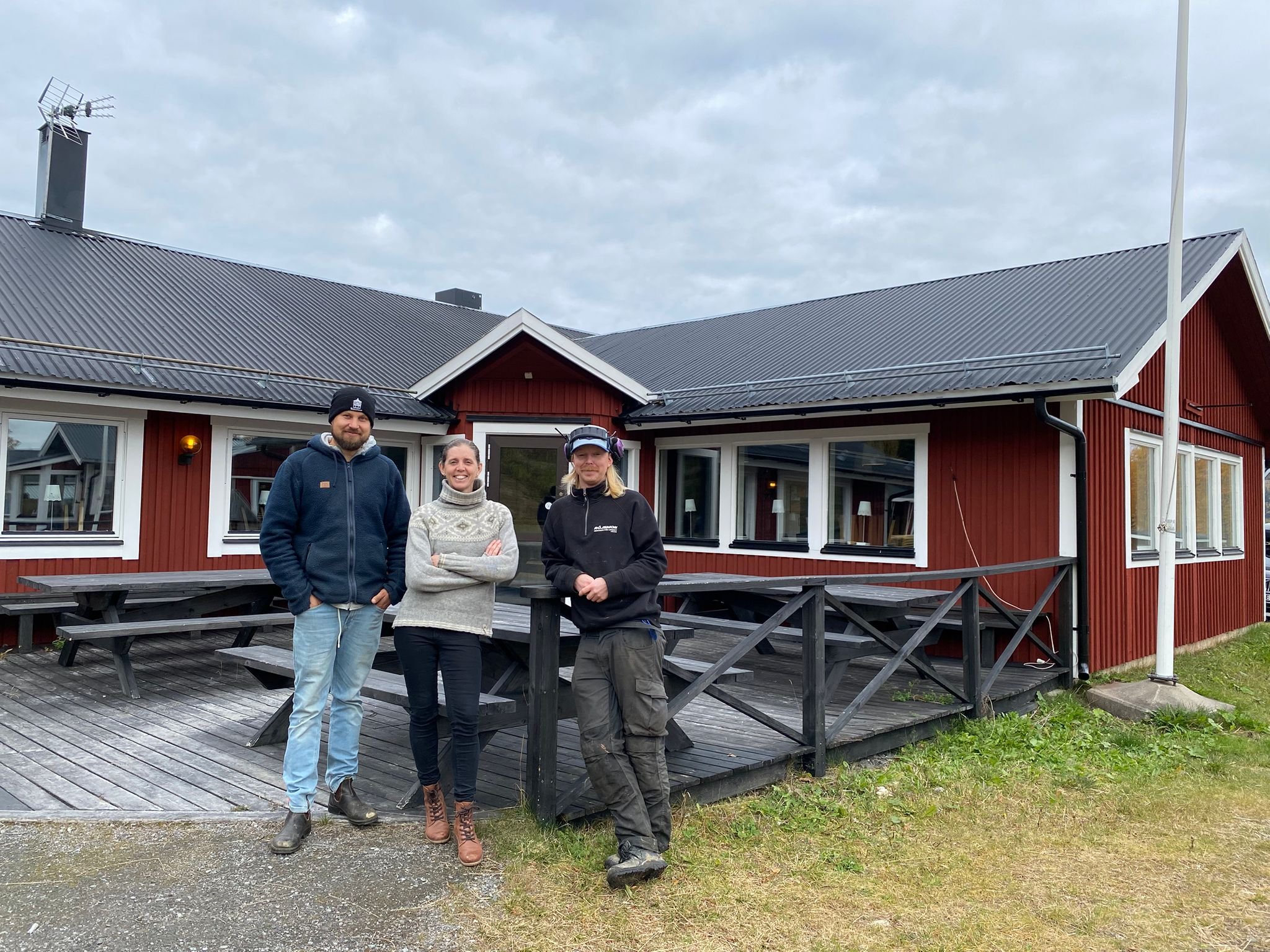 Tre personer står framför Gruvstugan i Huså: Johnny i mörkblå huvtröja, Karolina i en stickad tröja och en medarbetare i arbetskläder. De står på restaurangens uteservering av trä framför den röda träbyggnaden under en mulen himmel.