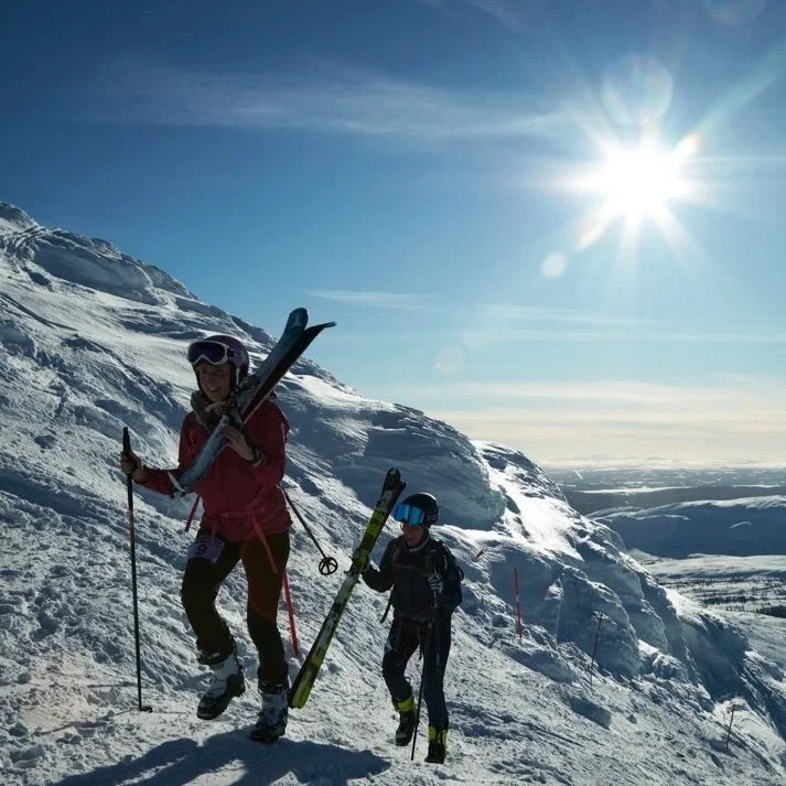 Två skidåkare som bär sina skidor på axlarna uppför en snöig bergstopp i strålande solsken. I bakgrunden syns ett vidsträckt vinterlandskap med fjäll och dalar.
