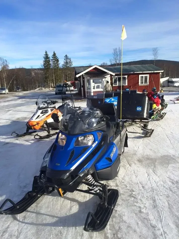 Flera snöskotrar parkerad i snön utanför en röd stuga i solsken.