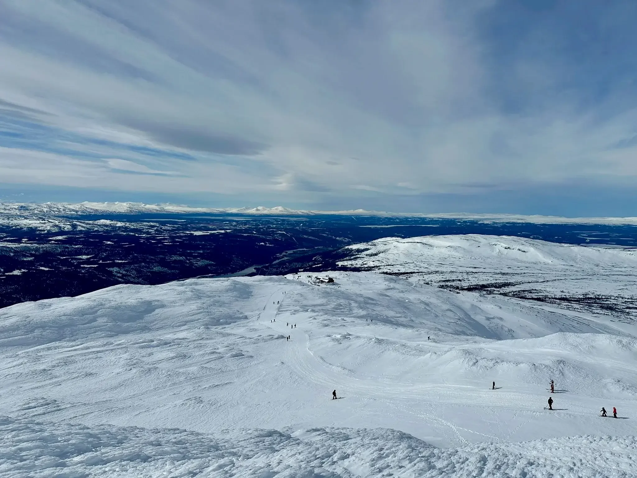 En konvoj med sex skidåkare på rad på en fjällled som sträcker sig genom ett vidsträckt, snötäckt fjällandskap under en mulen himmel.