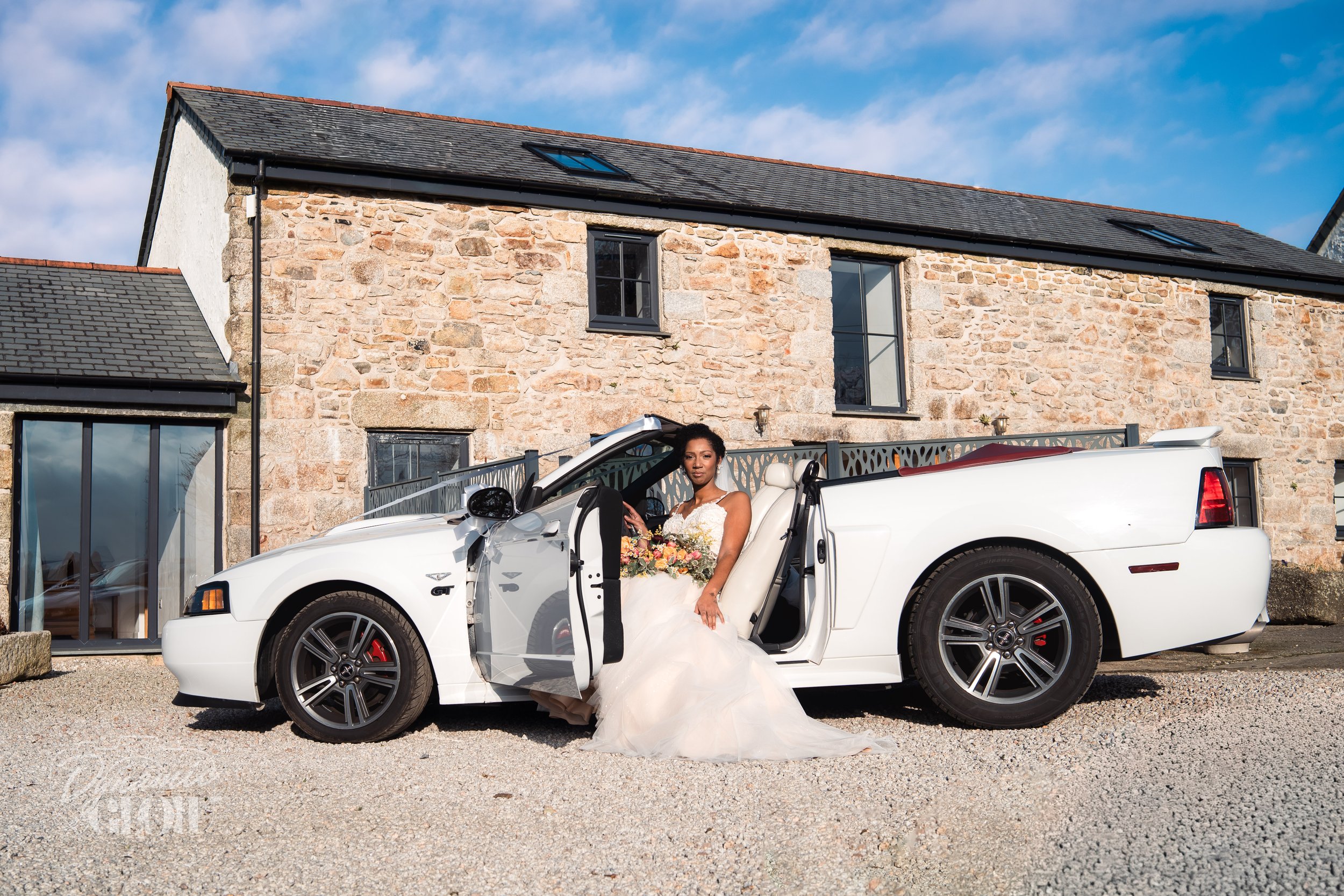 A bride in a wedding gown sitting in a white convertible car with the door open, holding a bouquet of flowers, in front of a stone house with a blue sky background.