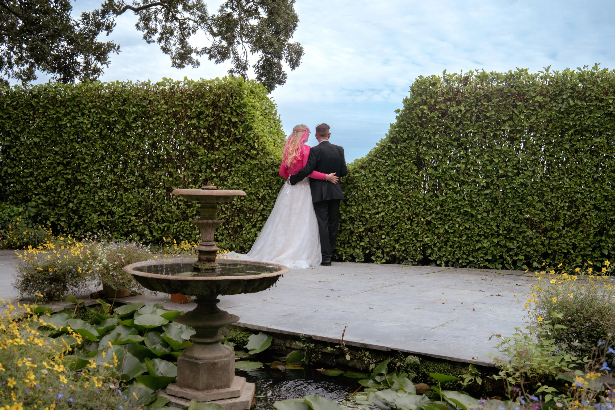 Bride and groom standing close together, facing away, with arms around each other in a garden with a hedge wall and fountain nearby.