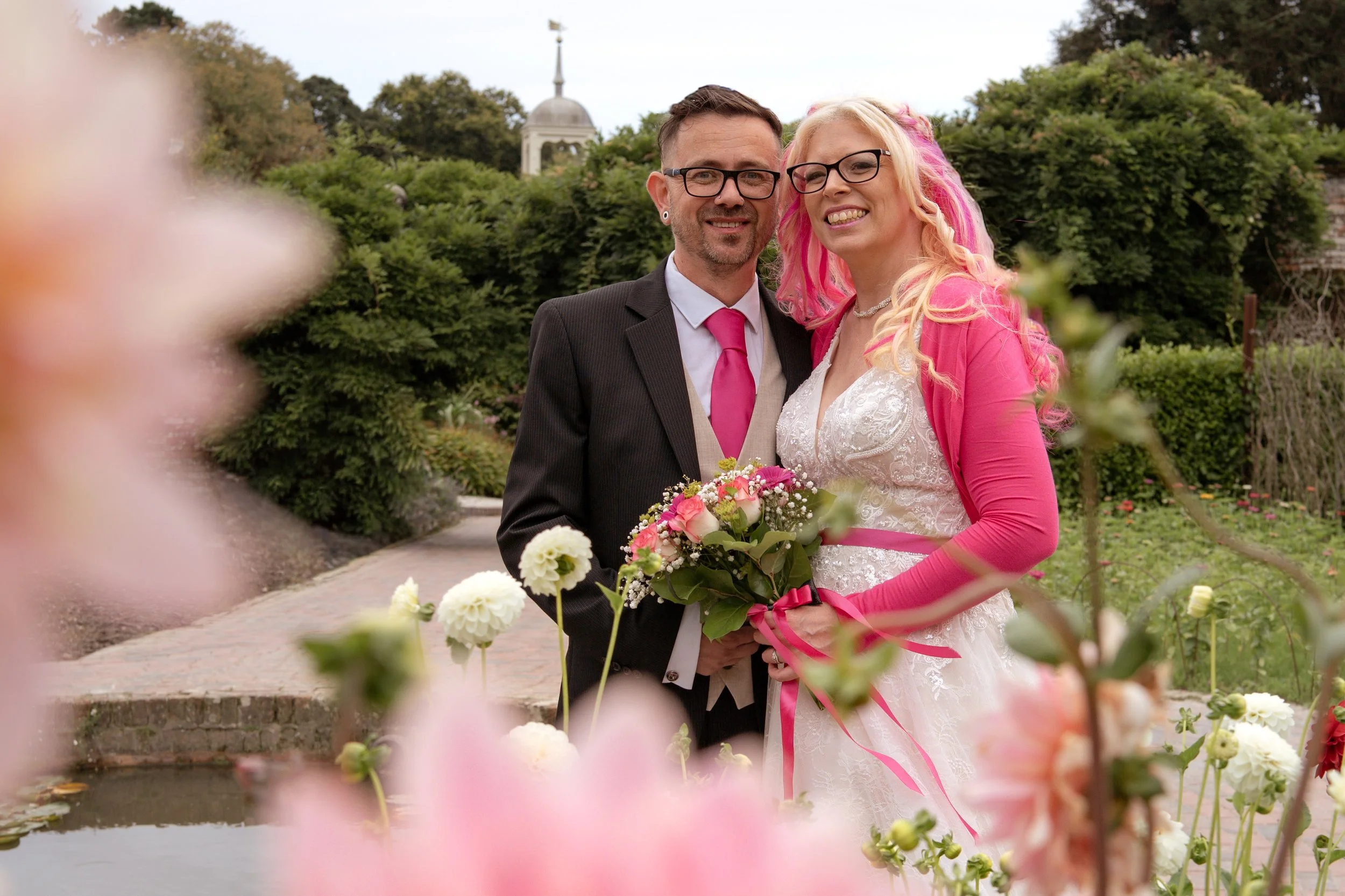 A happy couple at their wedding in a garden, with the bride holding a bouquet of flowers and wearing a pink cardigan over her white lace wedding dress, and the groom dressed in a suit with a pink tie, surrounded by pink and white flowers and greenery.