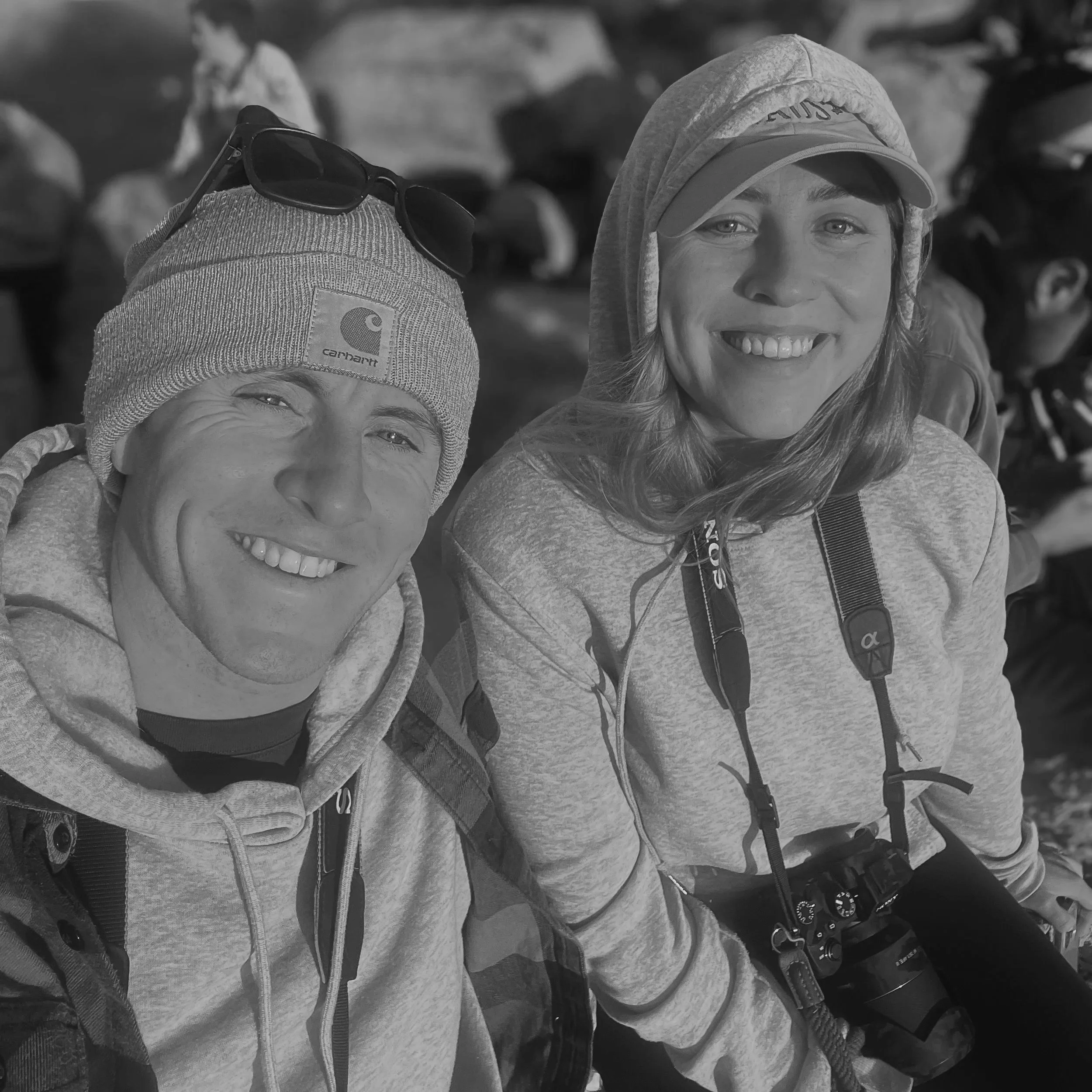 Close-up of two people wearing winter clothing, including hoodies and hats, with one person smiling, likely outdoors in a cold environment.