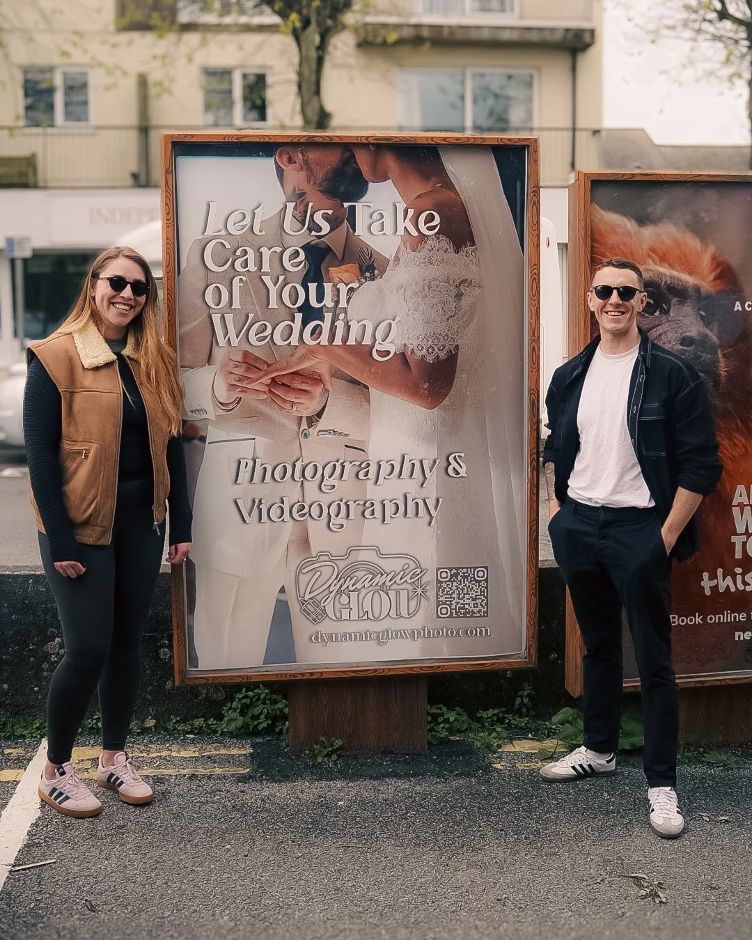 A woman and a man standing next to a sign promoting wedding photography and videography. The sign features a large image of a bride and groom and the text "Let Us Take Care of Your Wedding." The woman is wearing sunglasses, a black athletic outfit, and a brown vest, while the man is dressed in a white t-shirt, dark pants, plus a black jacket, and sunglasses.