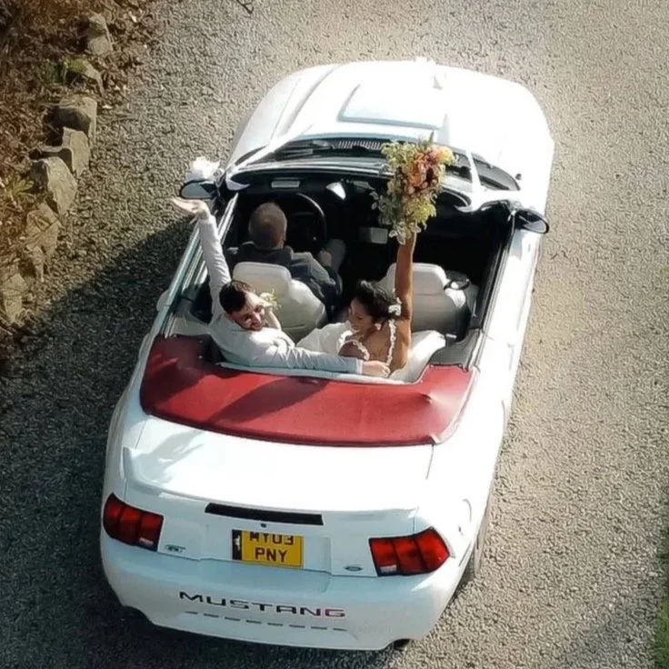 A convertible car with the top down, showing a bride and groom celebrating. The bride is holding a bouquet high in the air and the groom is smiling at her. The car is on a gravel road, with some rocks and grass visible at the sides.