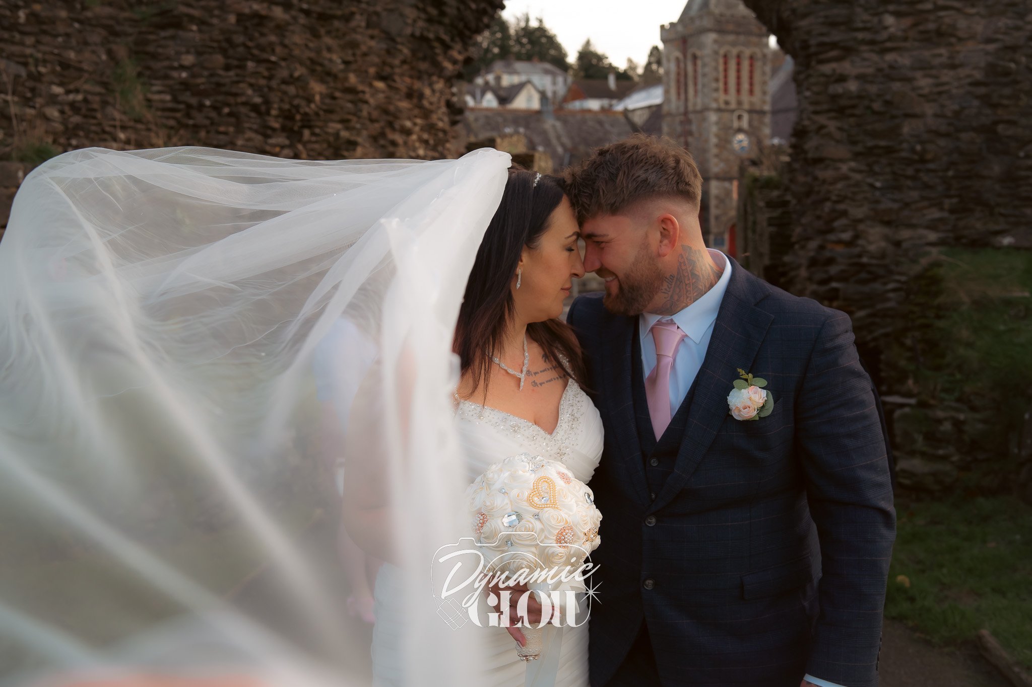 A bride and groom face each other closely, smiling, with their foreheads touching. The bride wears a wedding dress and veil, and the groom wears a suit with a boutonniere. The background features stone buildings and a church tower.