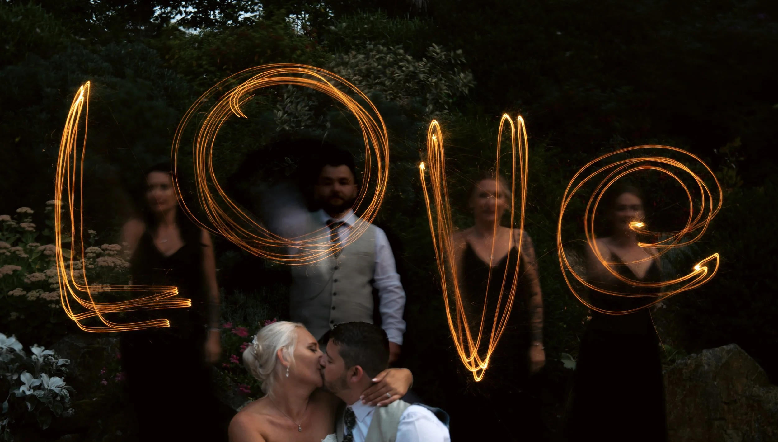 A couple is kissing in the foreground, surrounded by four people holding sparklers that spell out the word 'LOVE' against a dark outdoor background.