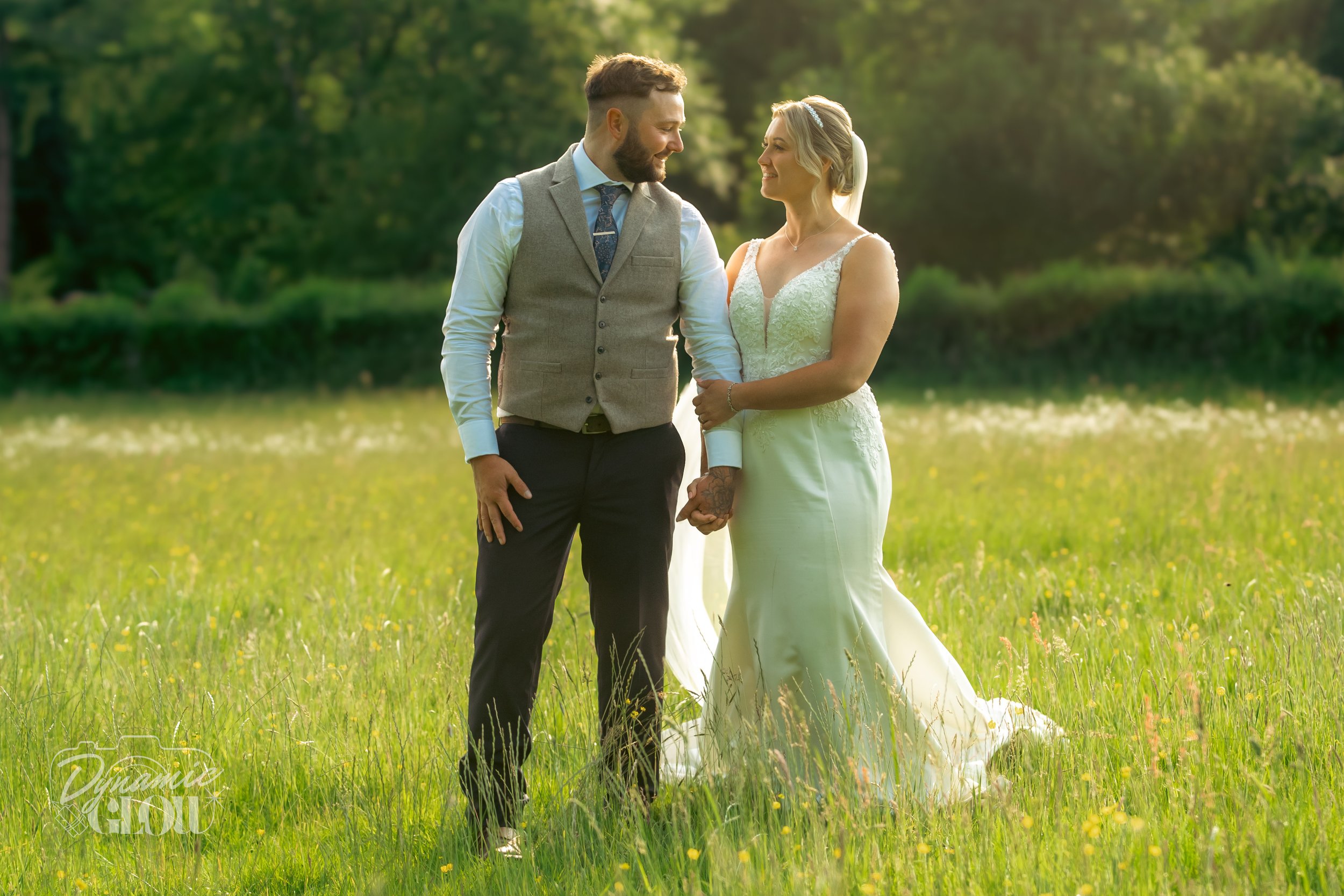 A bride and groom holding hands in a grassy field on their wedding day, with the groom wearing a vest and dress shirt, and the bride in a white gown with lace details, smiling at each other.