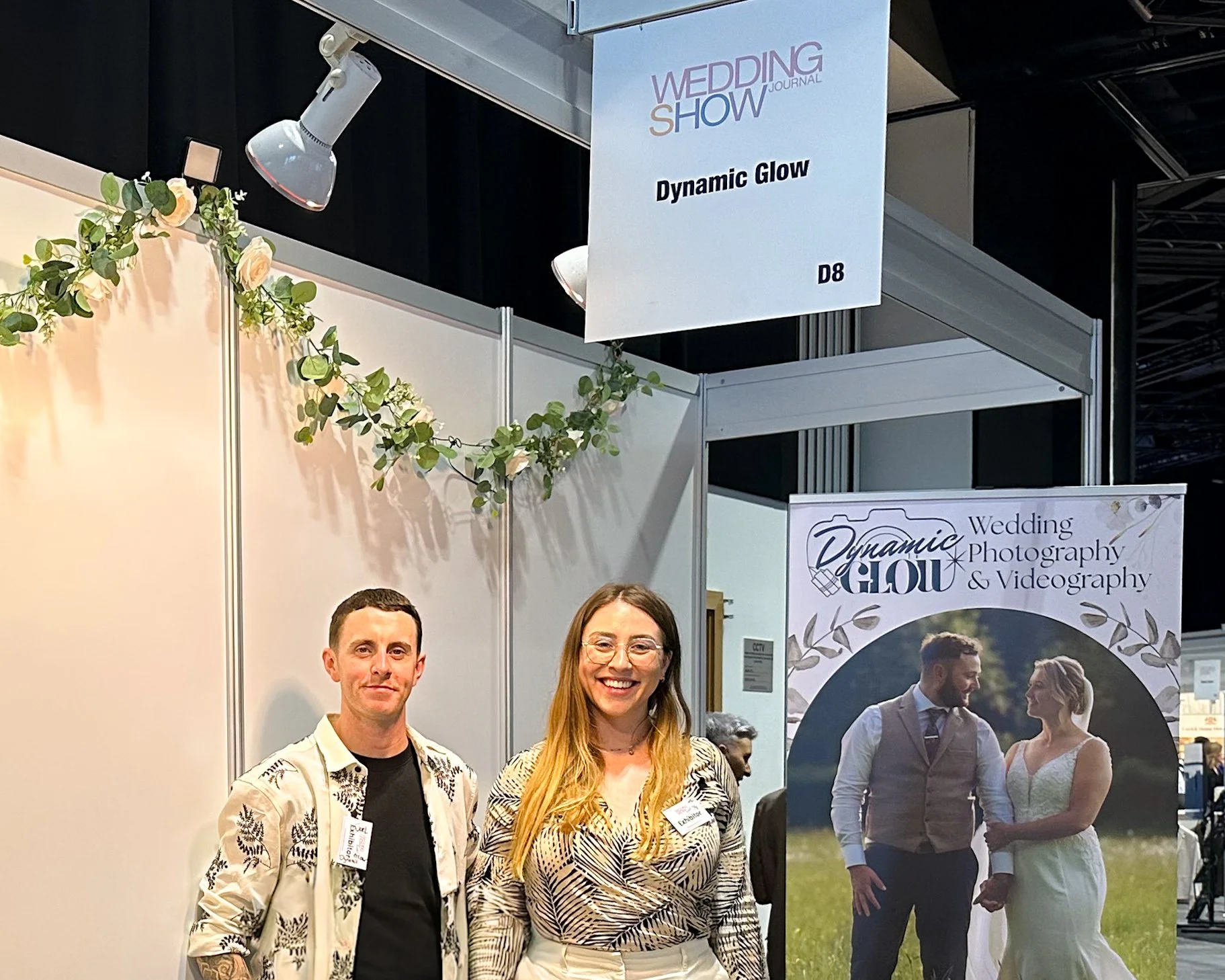 Two people standing in front of wedding photography display at a trade show. The man on the left is wearing a patterned blazer over a black shirt, and the woman on the right is wearing glasses and a patterned blouse. Behind them is a large poster with a wedding couple and text about wedding photography and videography. There is also a hanging sign that reads 'Wedding Show Journal' and 'Dynamic Glow' with booth number D8, decorated with greenery.