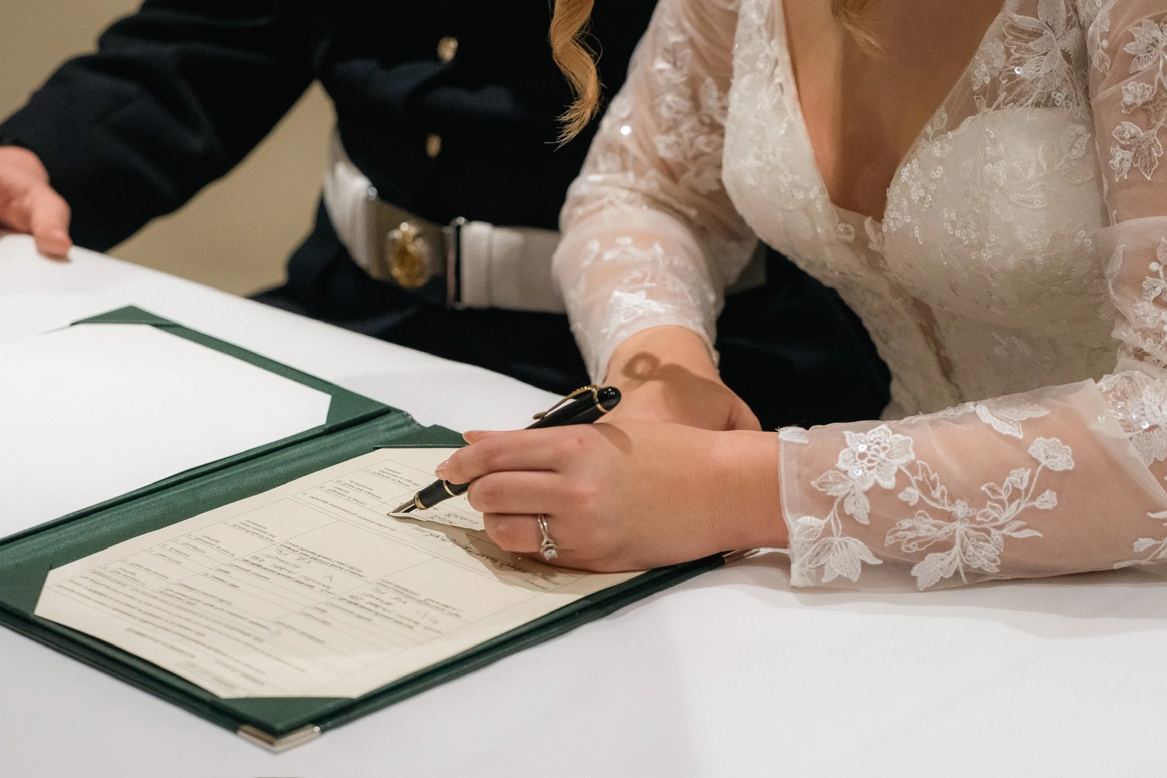 A woman in a lace wedding dress signing a document, with another person in a black uniform watching nearby.