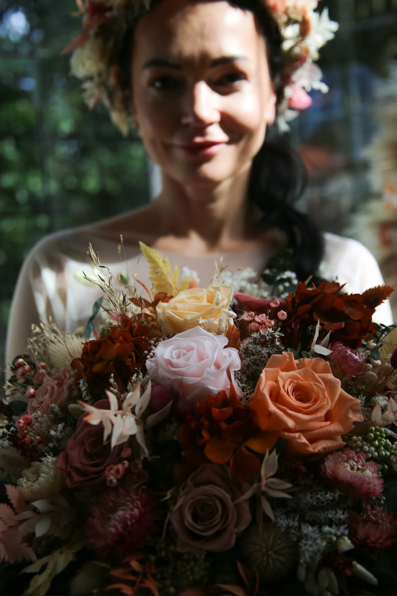 A woman with dark hair and a floral crown holding a large bouquet of multicolored roses and other flowers, smiling softly.