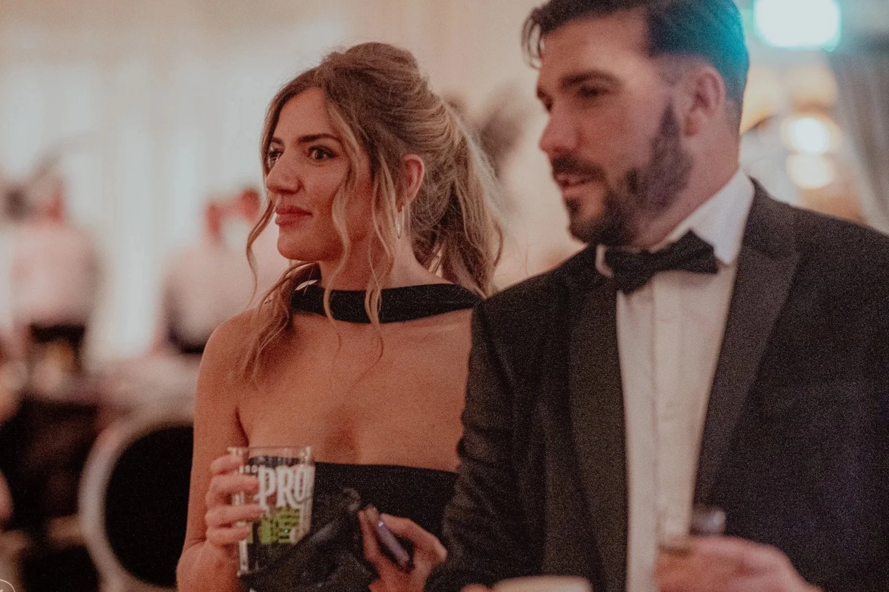 A woman in a black dress with a choker style neckline and a man in a tuxedo with a bowtie at a formal event, holding a drink and a phone, with blurred backgrounds of other guests.