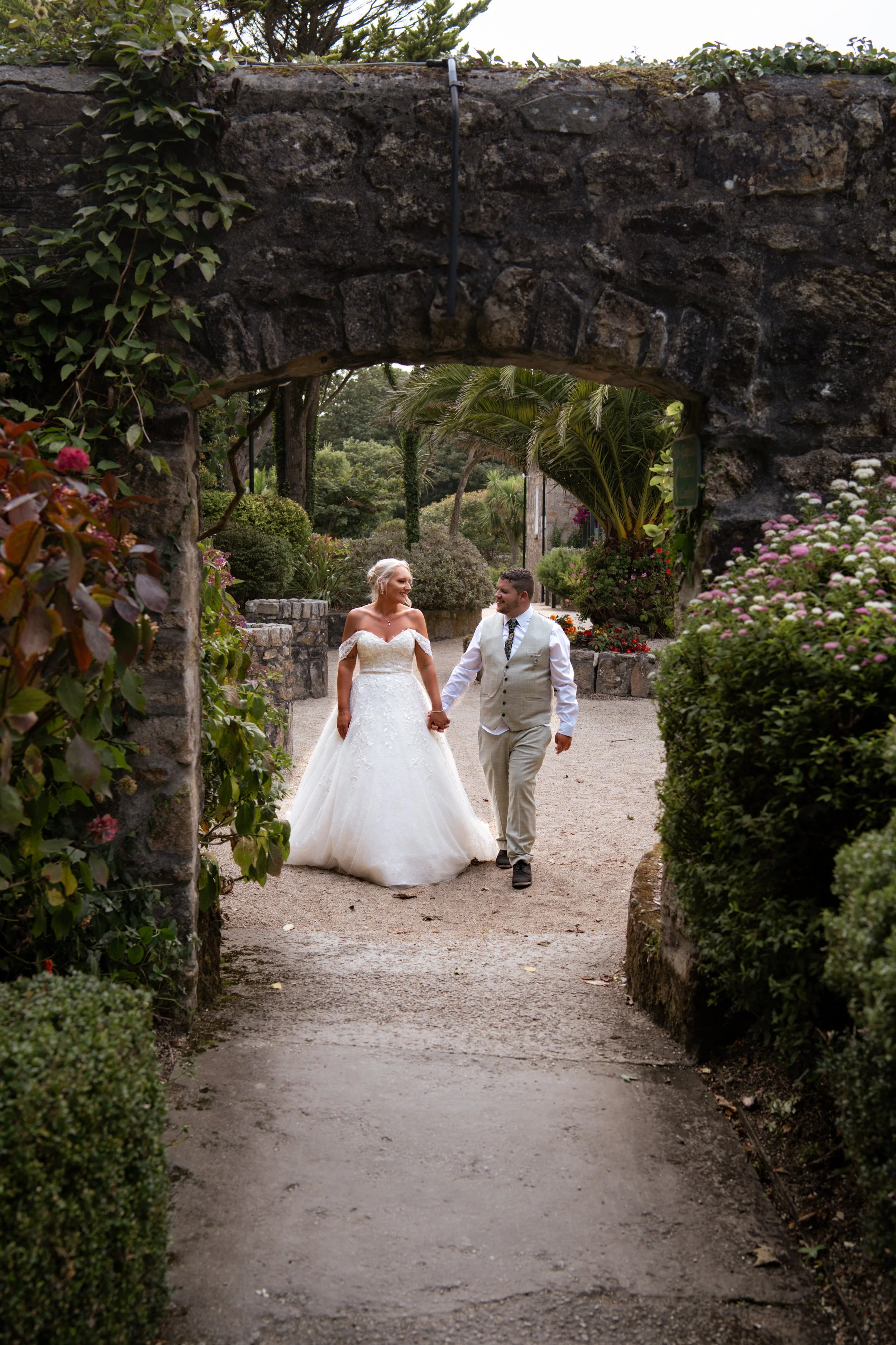 A bride and groom walking hand in hand through a garden archway, smiling at each other on their wedding day.