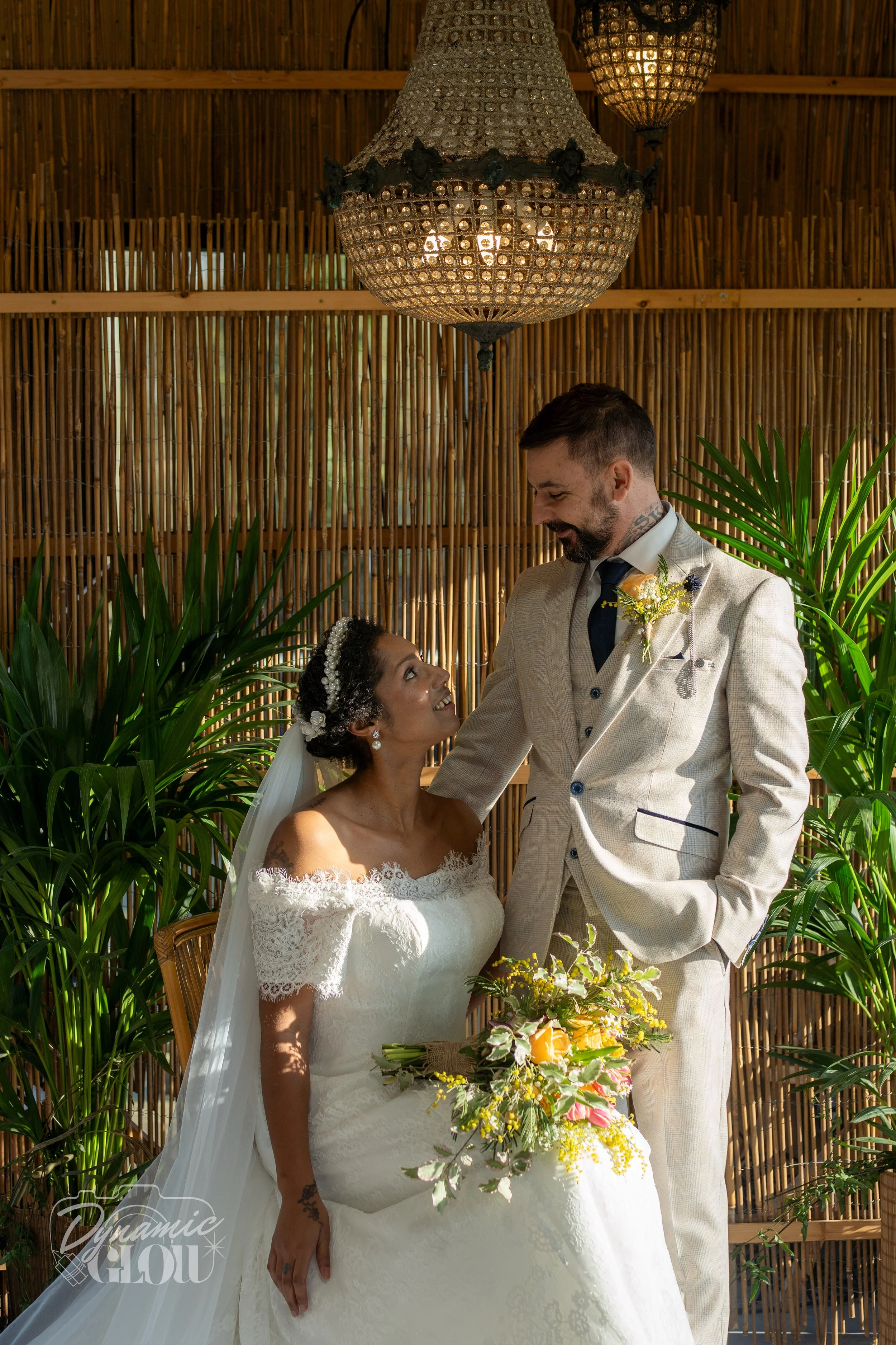 A bride and groom sharing a moment during their wedding ceremony, surrounded by lush green plants, with a bamboo wall background and a large ornate hanging lamp.