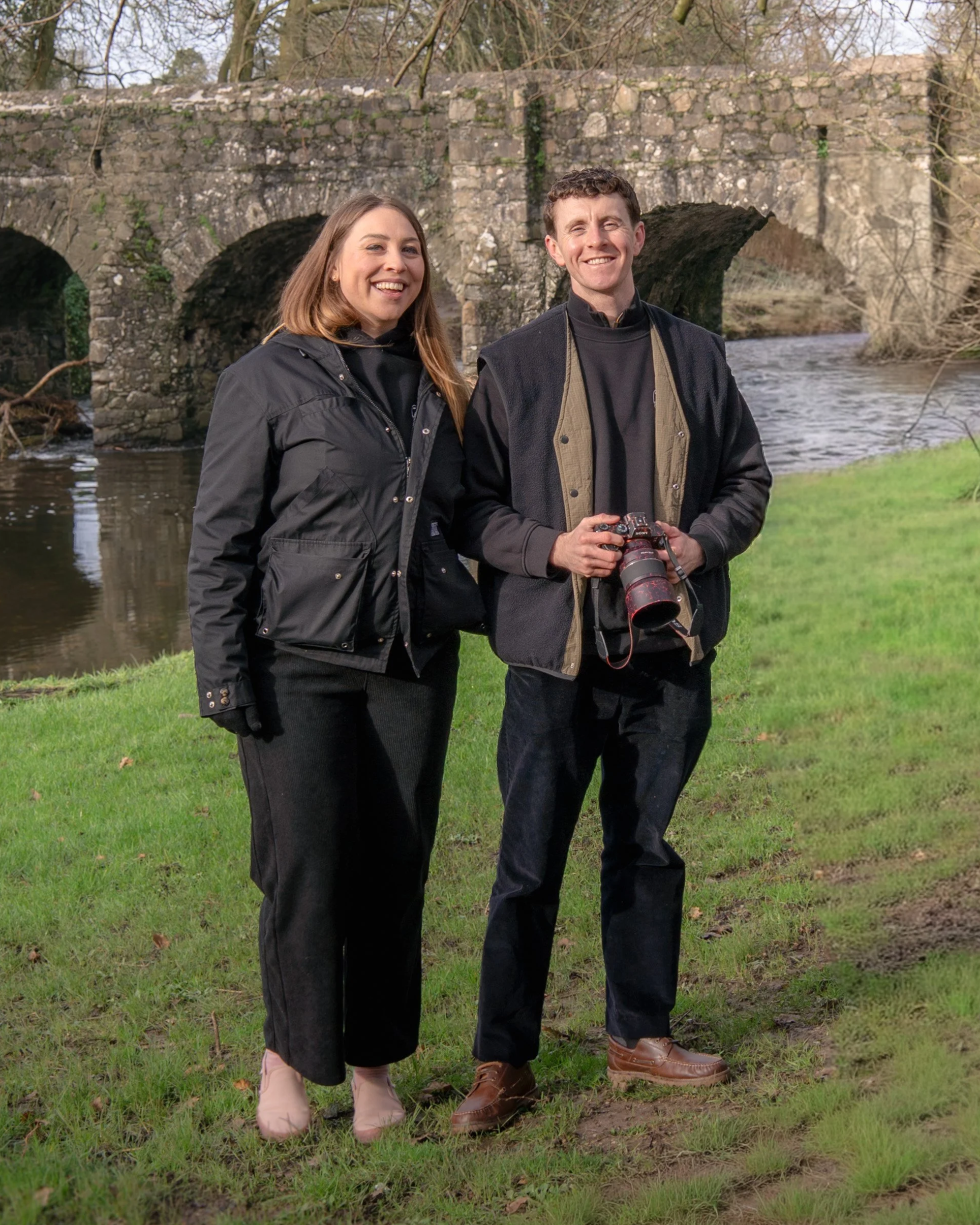 Two smiling people, a woman and a man, standing outdoors on grass near a stone bridge over a river, holding a camera, and dressed in outdoor clothing.