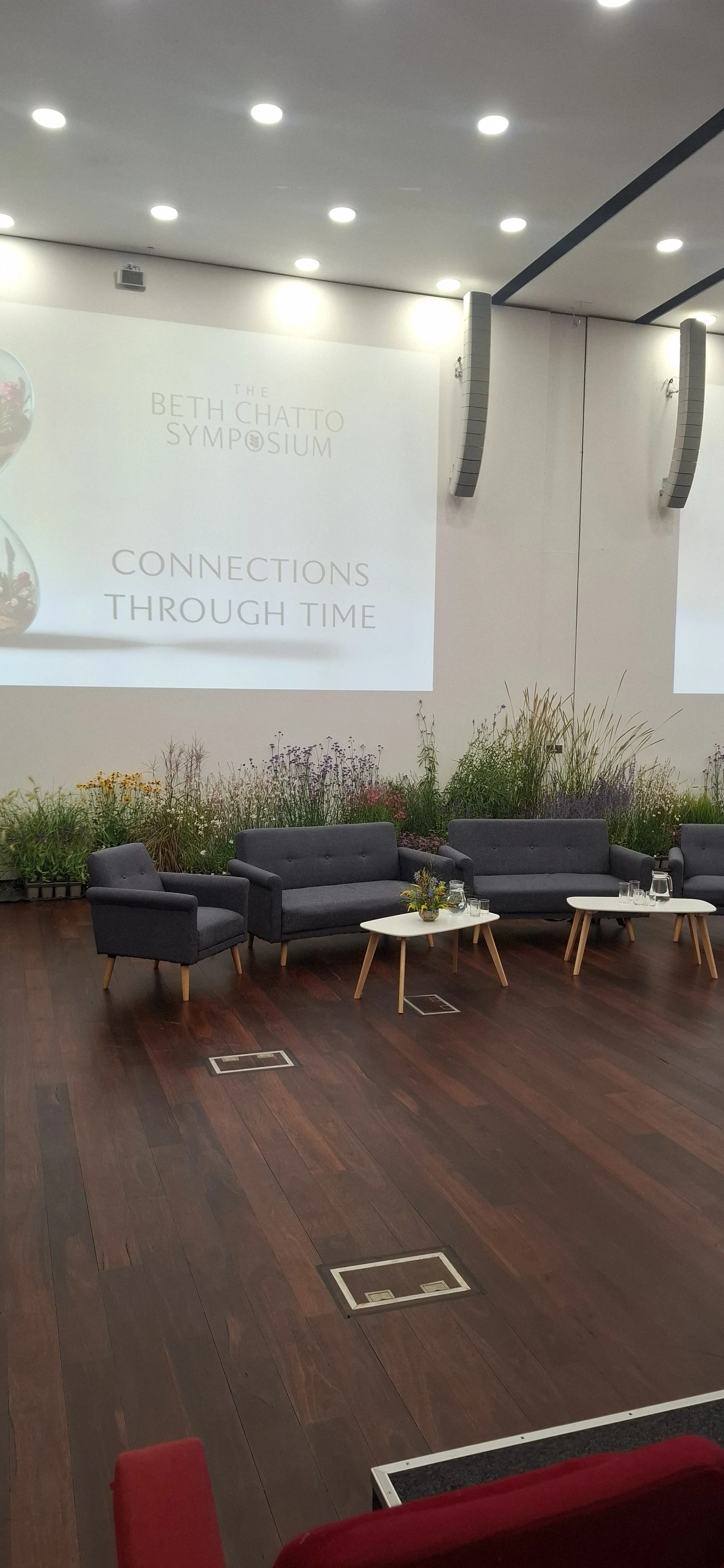 Modern symposium lounge with gray sofas, armchairs, coffee tables, flowers, and a projected sign reading “The Beth Chatto Symposium: Connections Through Time.”