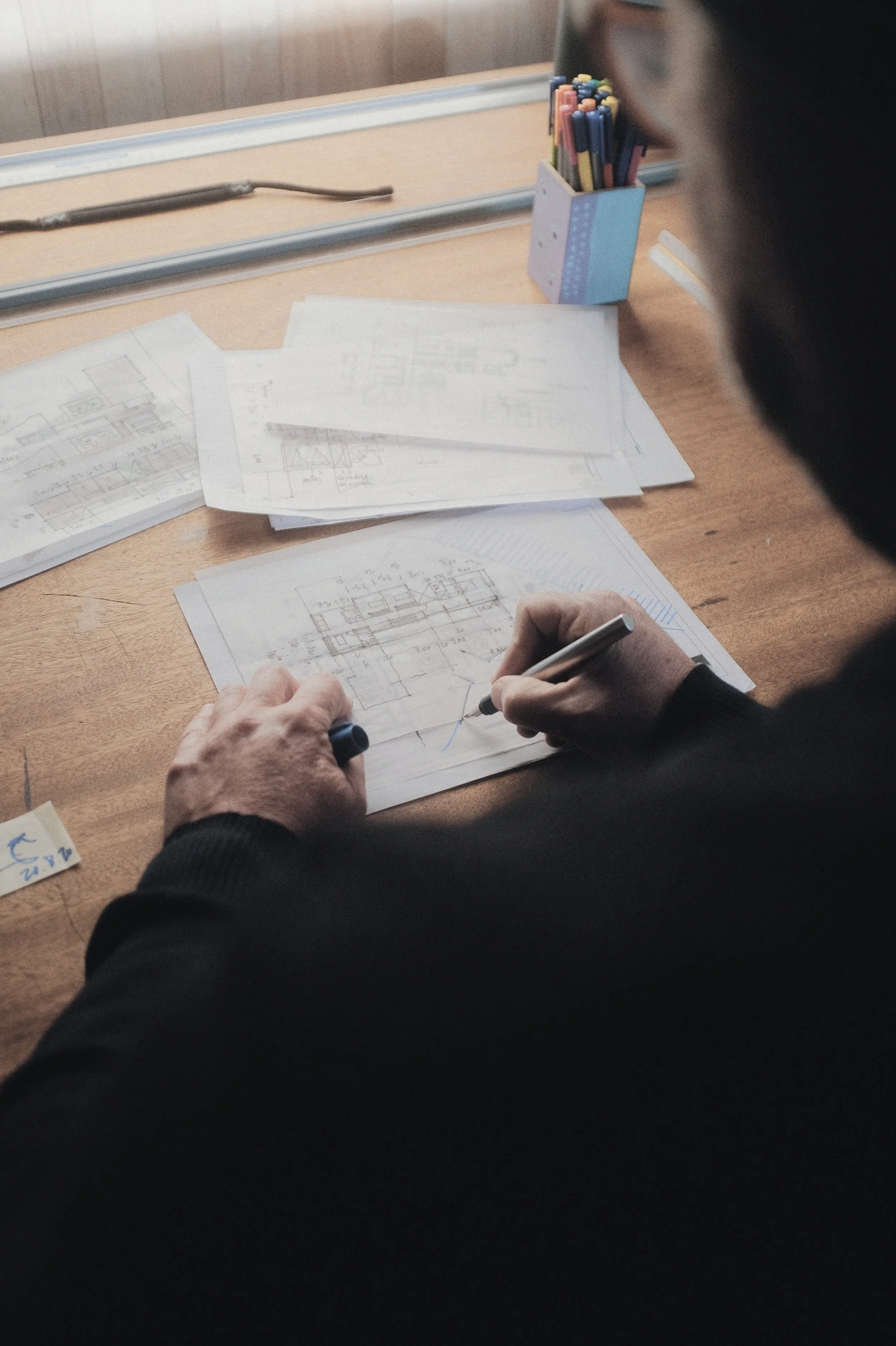 A person is working on architectural blueprints at a wooden desk, with a pen in hand, and a container of colored pencils nearby.