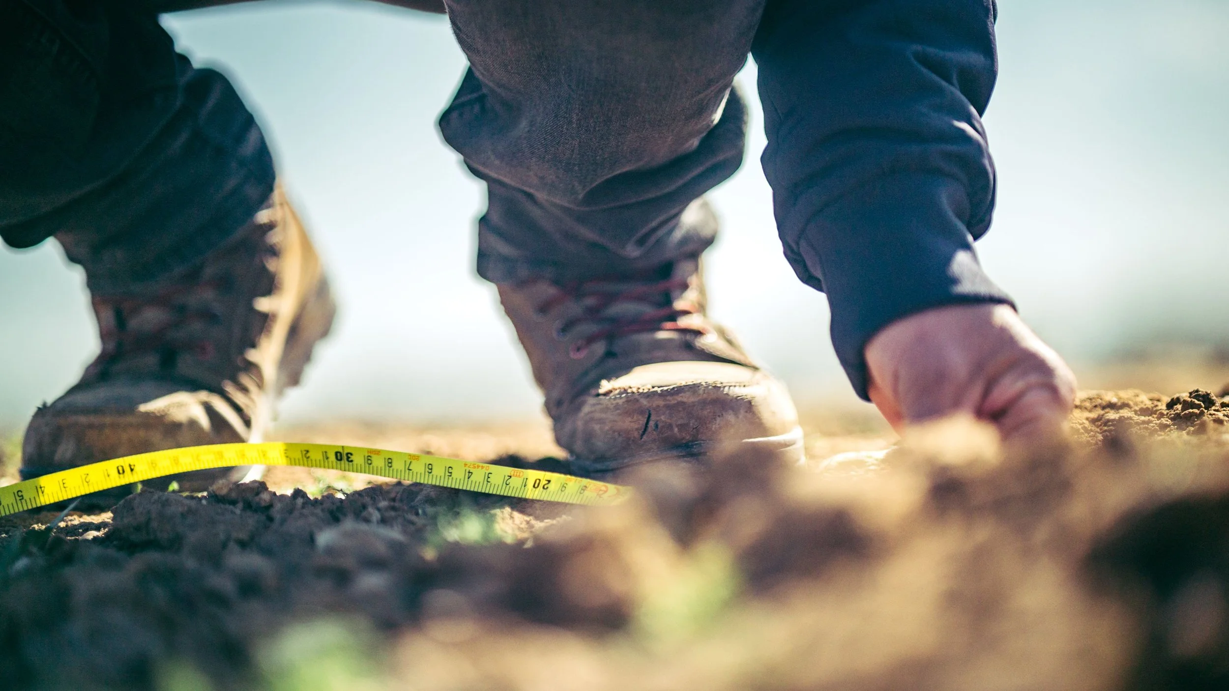 Close-up of a person wearing brown boots and black pants kneeling on soil, using a measuring tape to measure the ground outdoors.