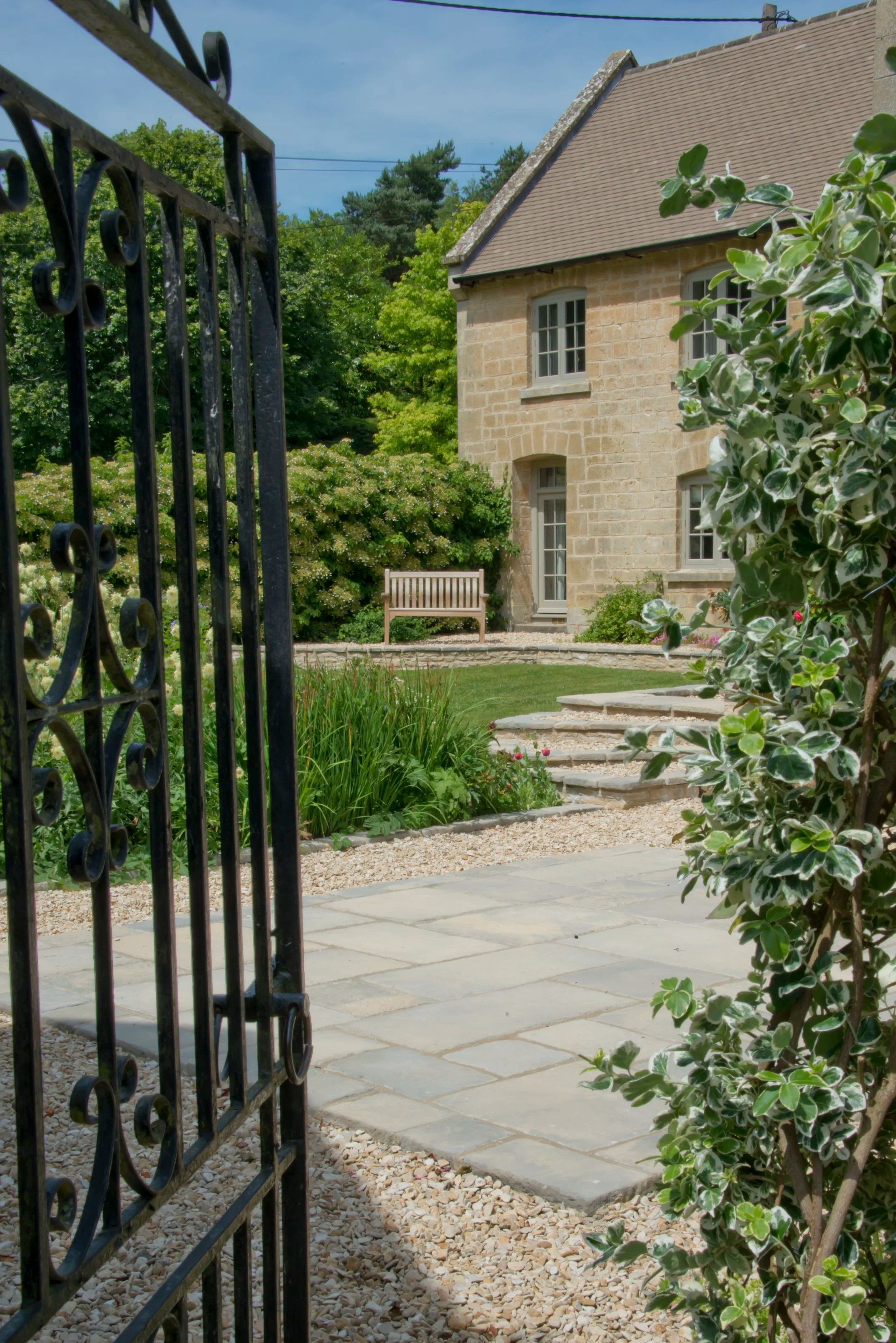 View of a garden with a stone house, pathway, bench, lush greenery, and plants, seen through an open wrought iron gate.