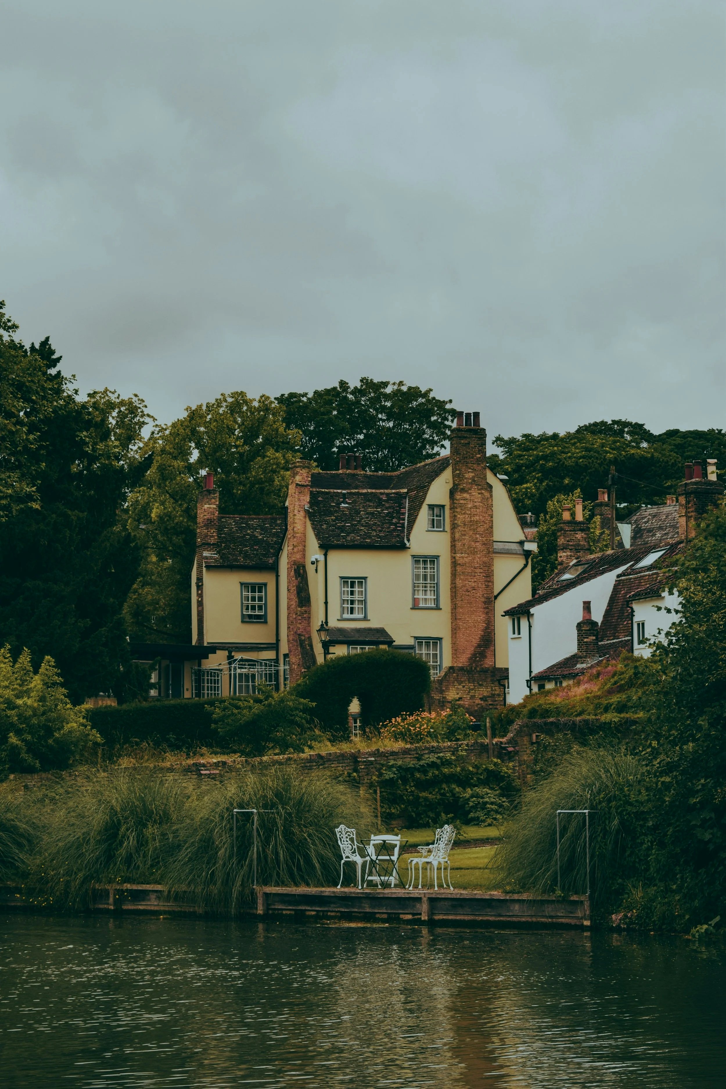 A picturesque scene of a traditional cottage with a garden and patio furniture next to a body of water under a cloudy sky.