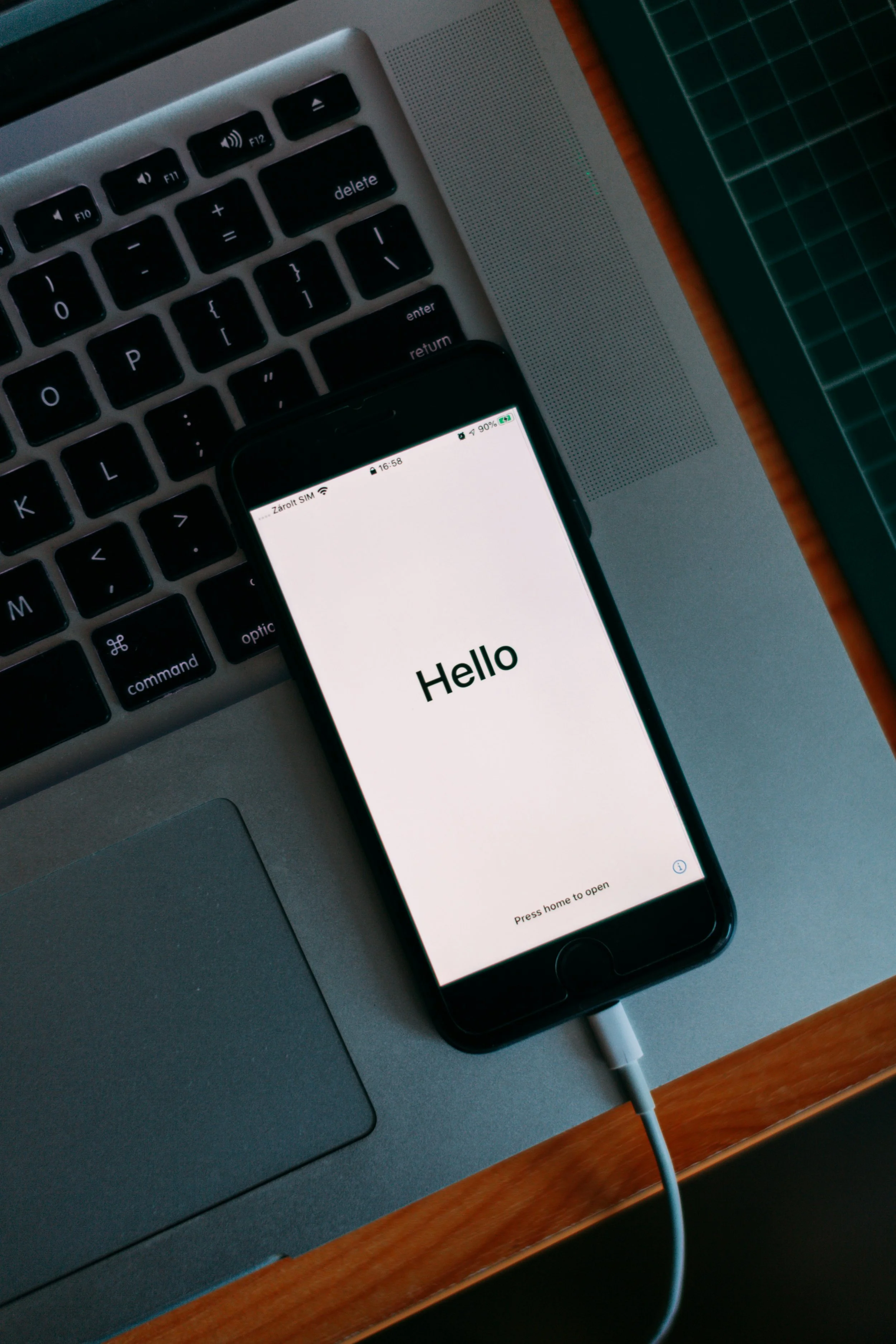 Close-up of a laptop keyboard, a smartphone displaying a 'Hello' message, and a charging cable connected to the phone, on a wooden desk.