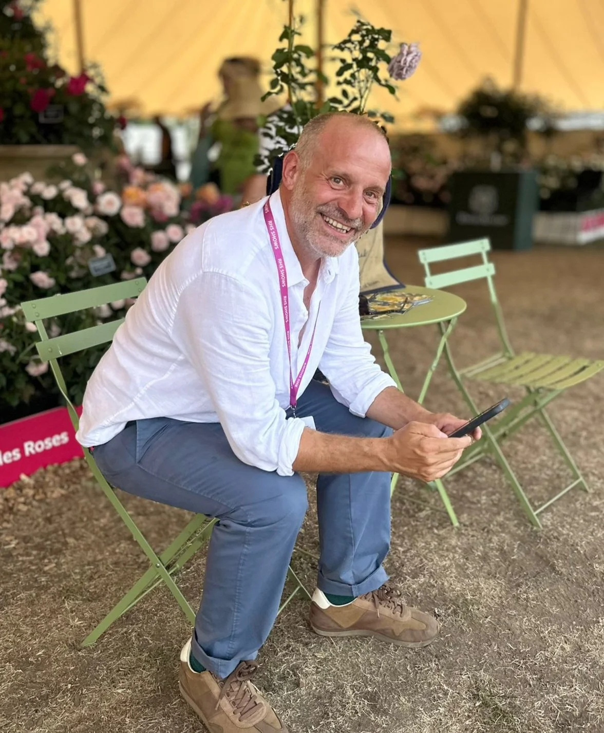 A smiling man in a white shirt and blue pants sitting on a green chair at a garden show, holding a phone, with pink flowers and garden displays in the background.