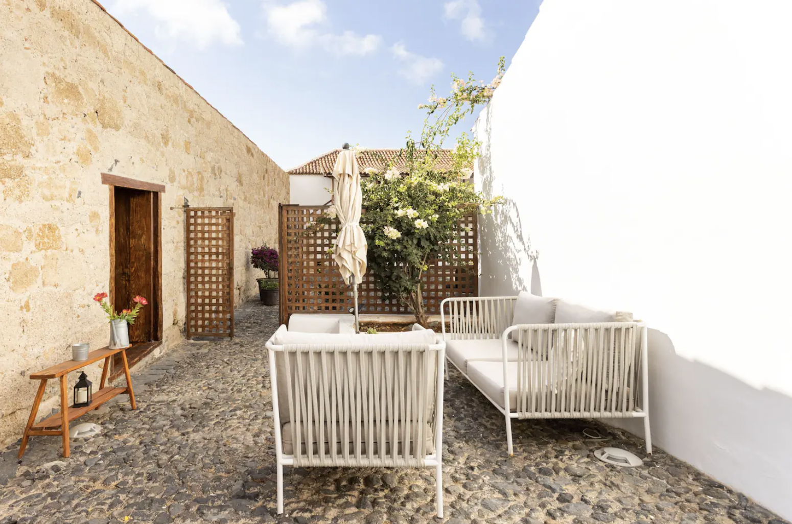 Cobblestone patio with white outdoor furniture, wooden trellises, a closed umbrella, potted plants, and a rustic wall.