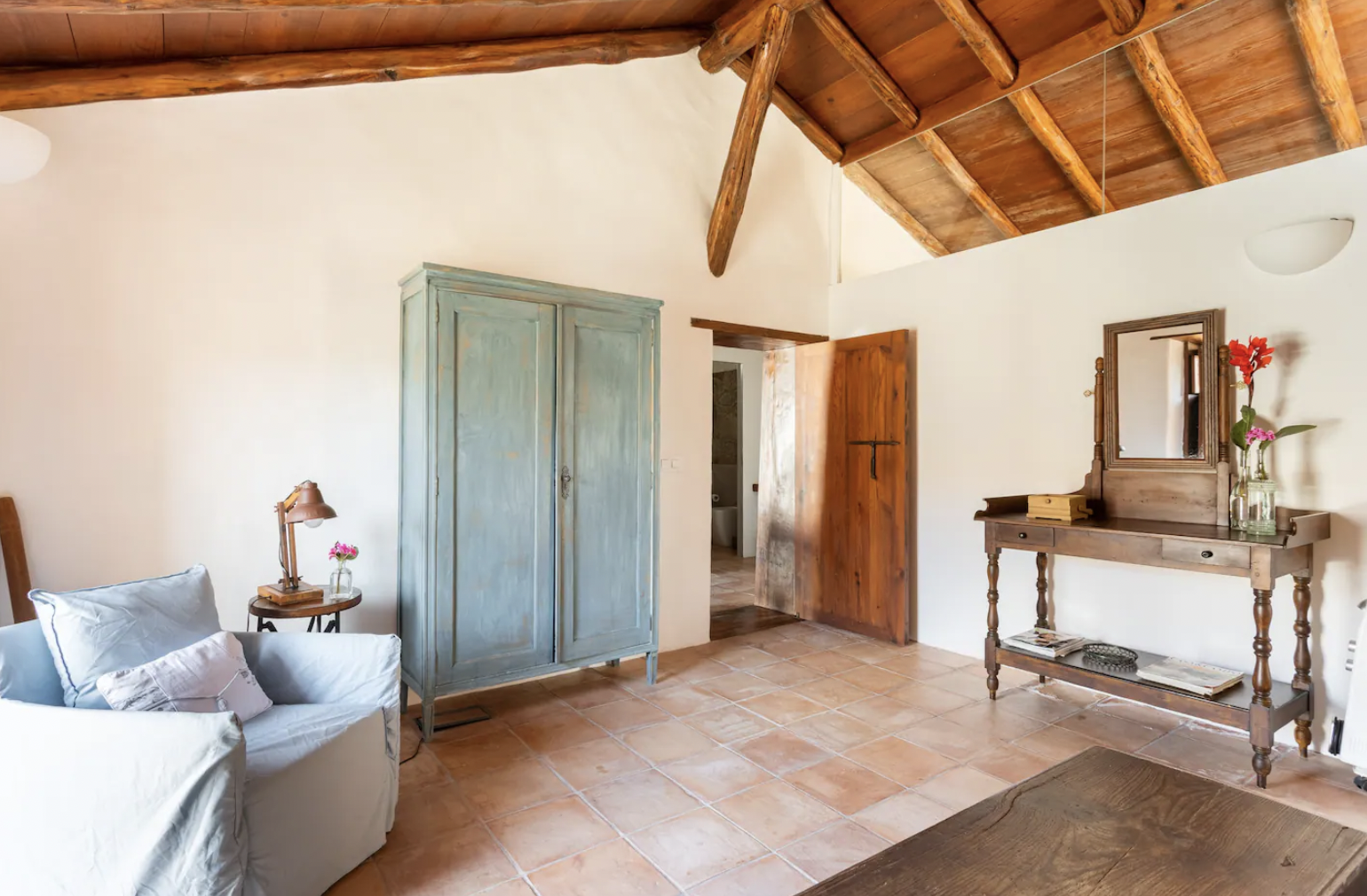 Rustic living room with wooden ceiling beams, terracotta floor, blue wardrobe, and vintage furniture.