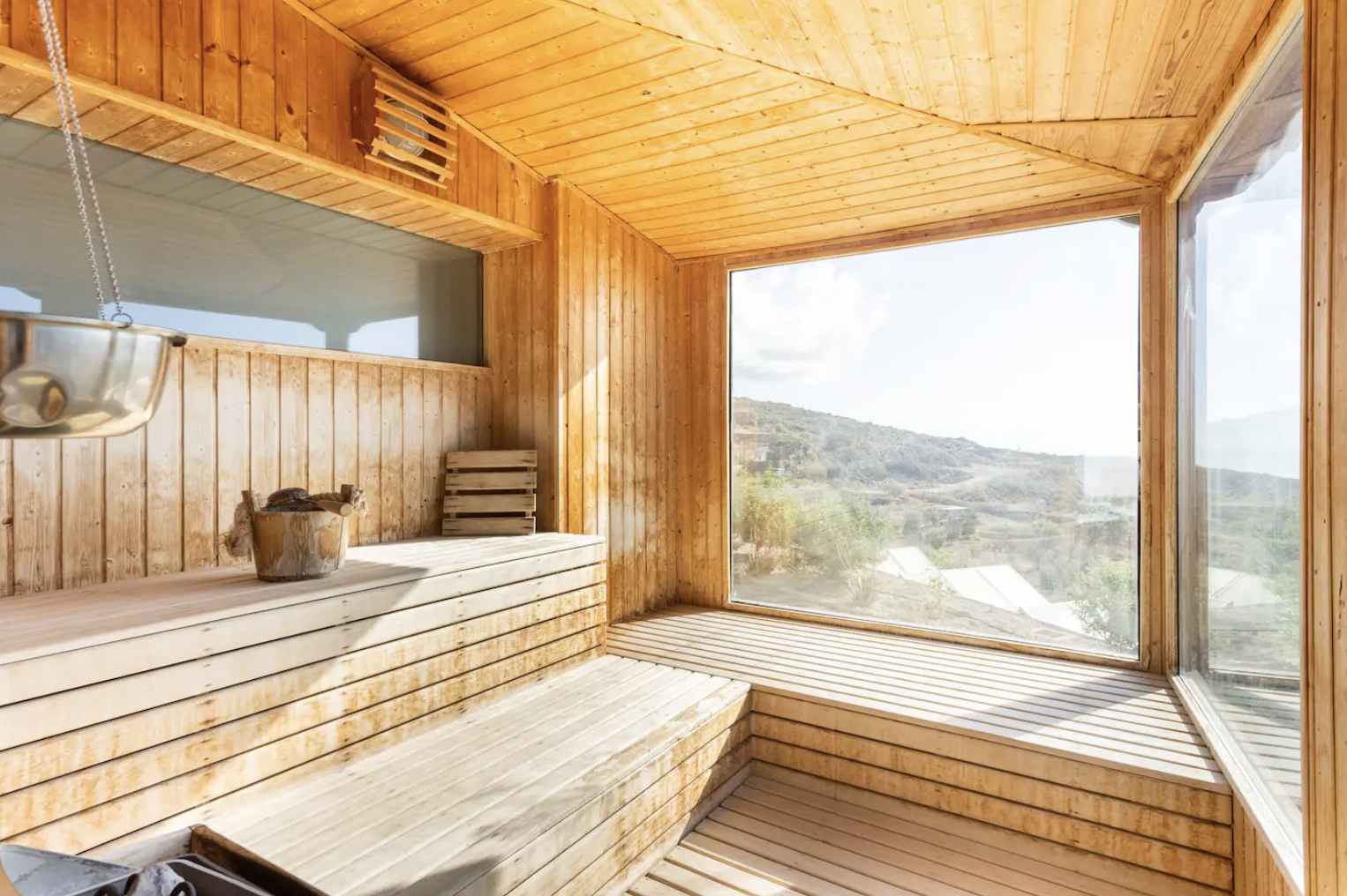Wooden sauna interior with large window overlooking scenic landscape, bench seating, and a bucket with a ladle.