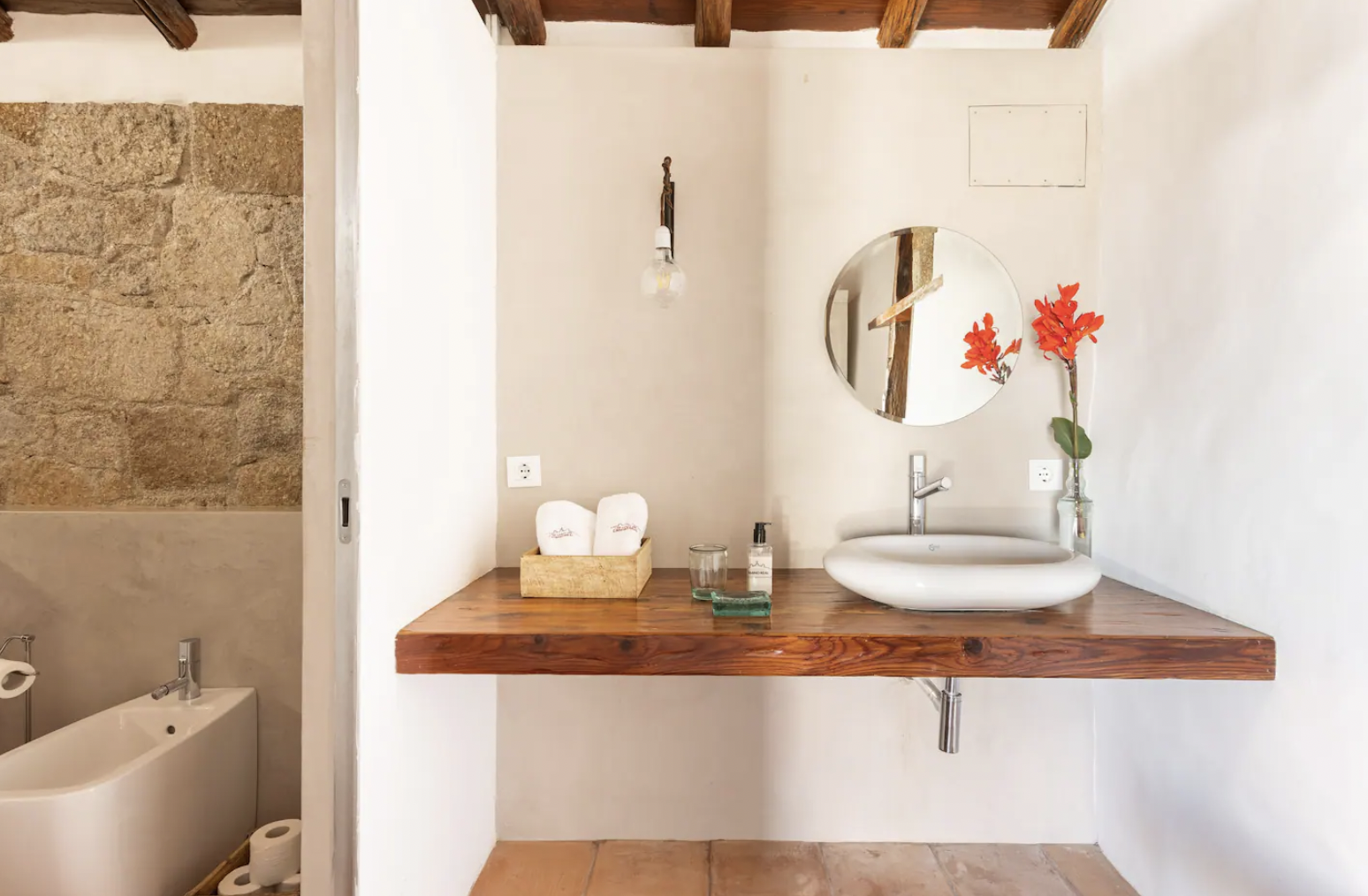Minimalist bathroom with a wooden countertop featuring a round sink, a circular mirror, a vase with red flowers, and beige walls with a stone accent. A bidet and a modern faucet are also visible.