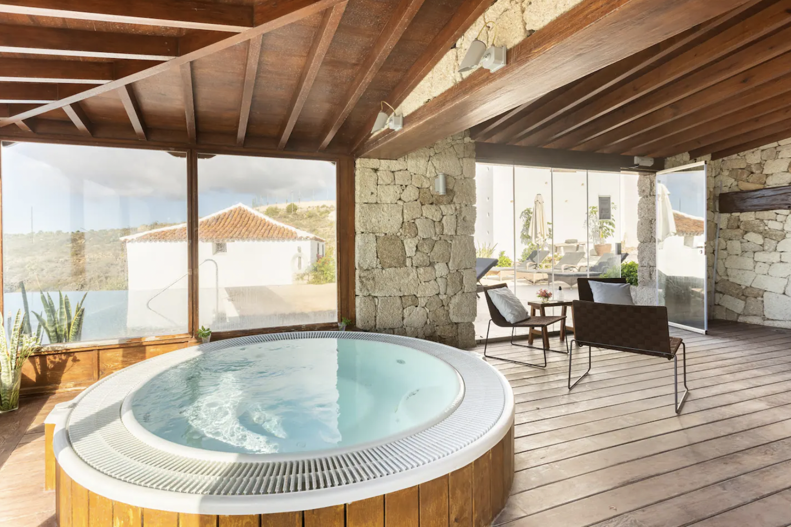 Indoor space with a hot tub, wooden ceiling and floor, large windows, stone walls, and seating area with chairs and a table.
