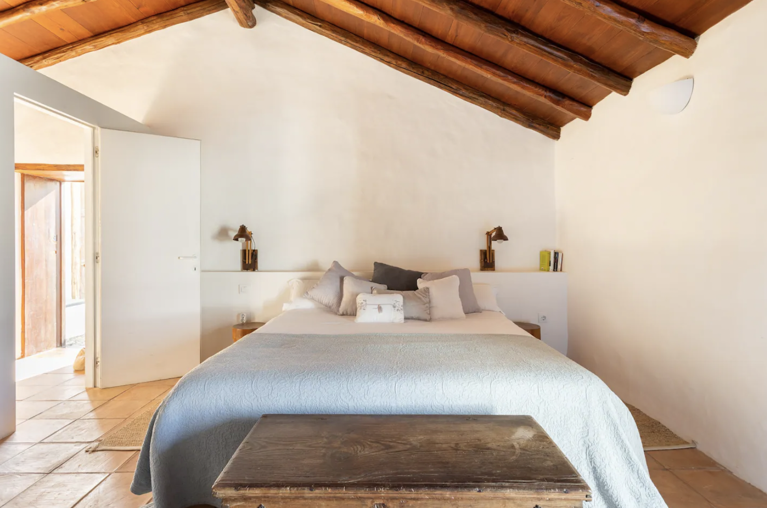 Cozy bedroom with a wooden ceiling, white walls, and a large bed covered in light bedding. Two lamps are attached to the wall behind the bed, and a wooden chest sits at the foot of the bed. The room has an open door leading to a bright space.