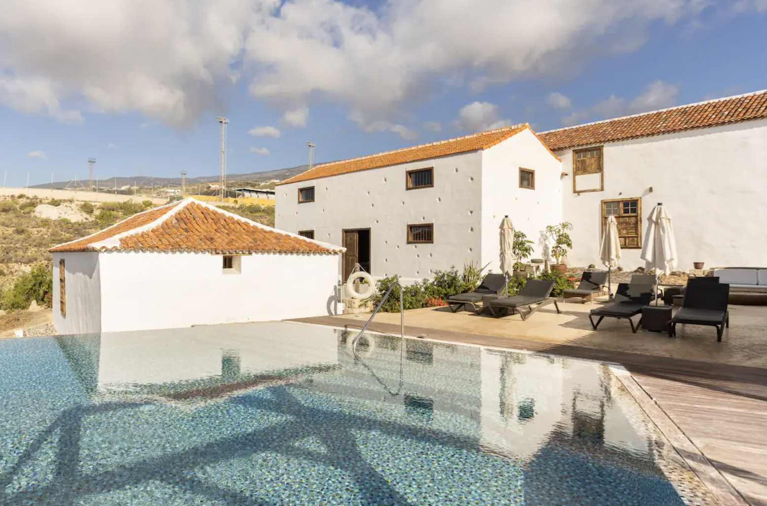 White stucco house with red-tiled roofs, an outdoor pool, sun loungers, and umbrellas surrounded by a patio, set in a rural landscape under a blue sky with clouds.