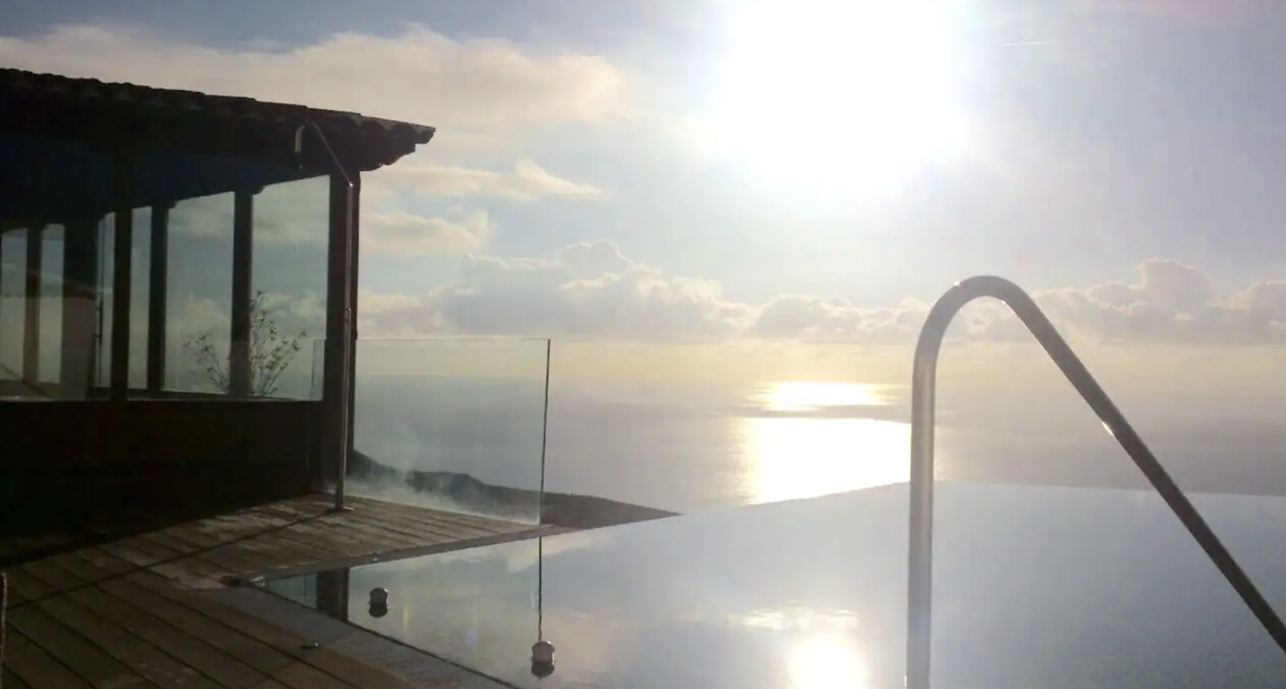 Infinity pool overlooking ocean with sun reflecting on water, glass railing, and adjacent building with large windows.