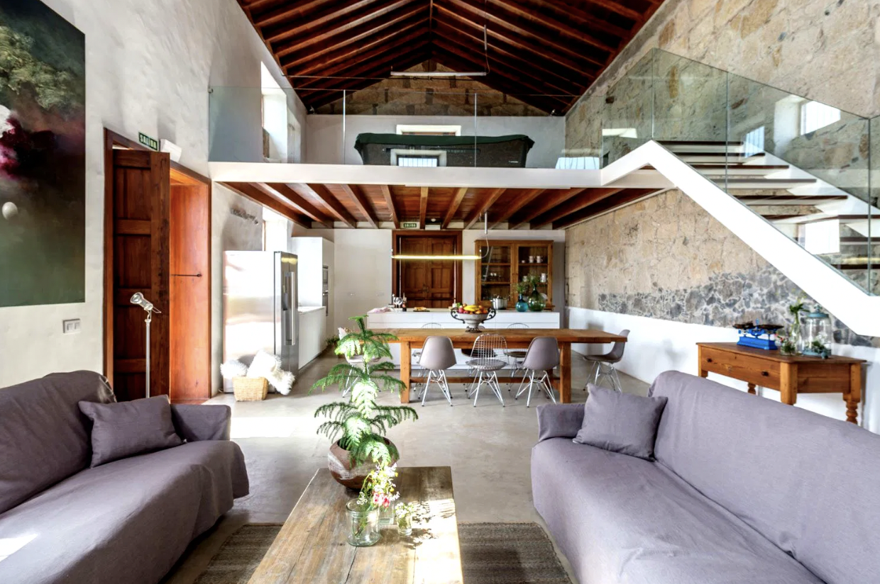 Modern loft interior with high ceilings, exposed beams, and stone walls. Features two gray sofas, a wooden coffee table with plants, a dining area with a long table and chairs, and an upper level with glass railings. Natural light enters through large windows.
