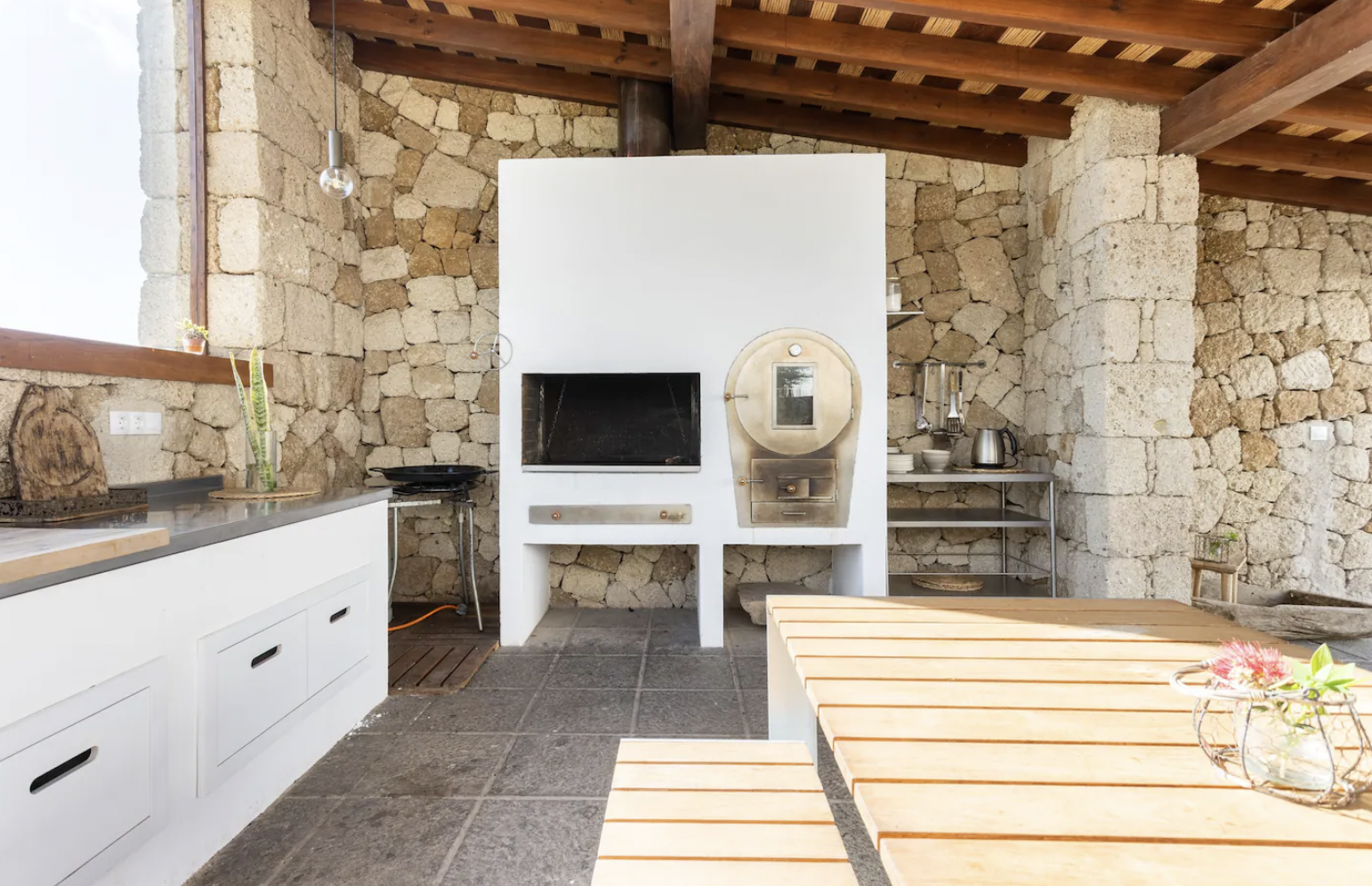 Outdoor kitchen with white cabinetry, stone walls, wood-burning oven, grill, potted plants, wood and metal table, and stone tiles.