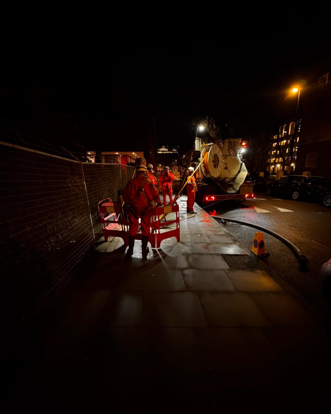 Nighttime scene with construction workers in red safety gear working on a sidewalk, using a large vacuum truck nearby, illuminated by streetlights.