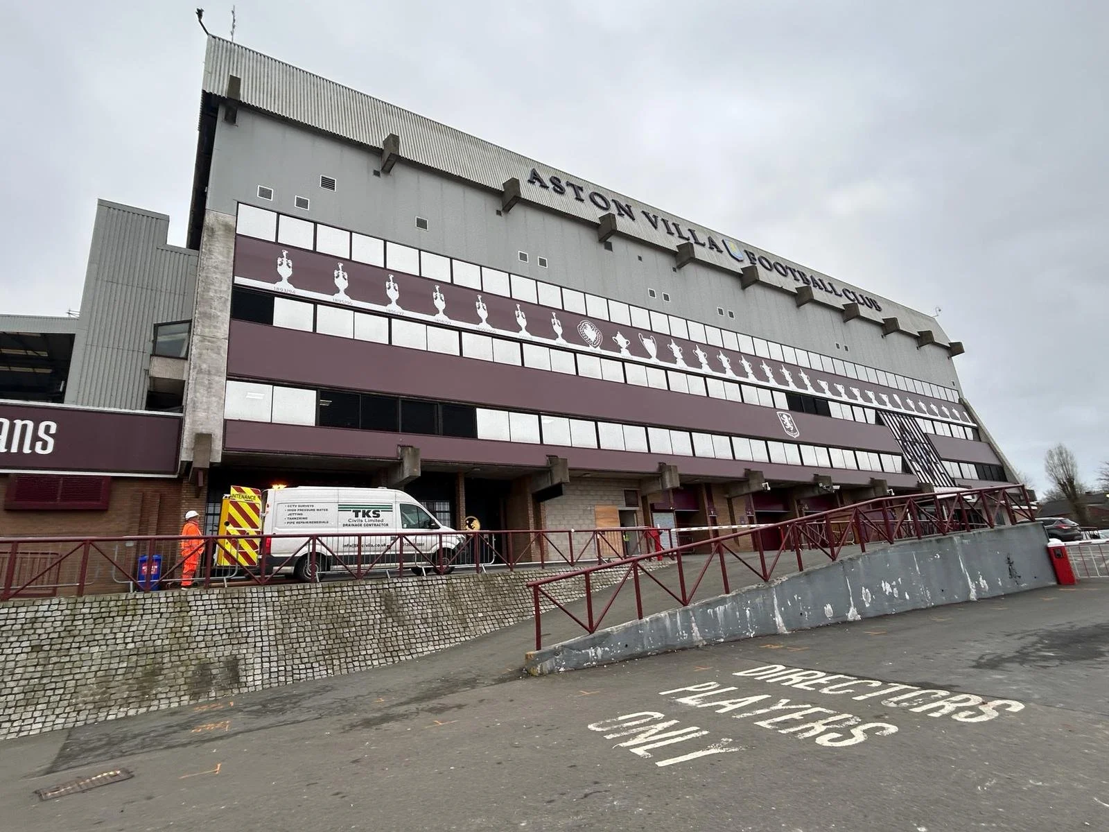 Exterior of Aston Villa Football Club stadium on a cloudy day, with a white van, a person in orange work clothing, and warning signs on the ground.