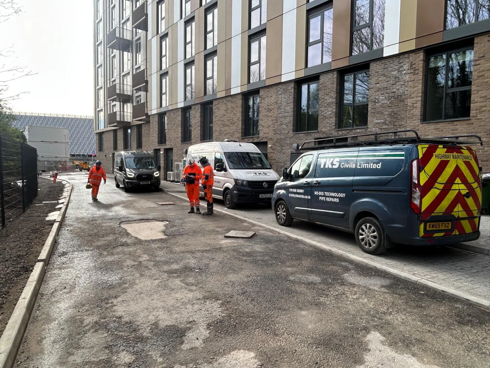 Construction workers in orange uniforms working on a street in front of a modern multi-story building, with maintenance vans parked nearby.