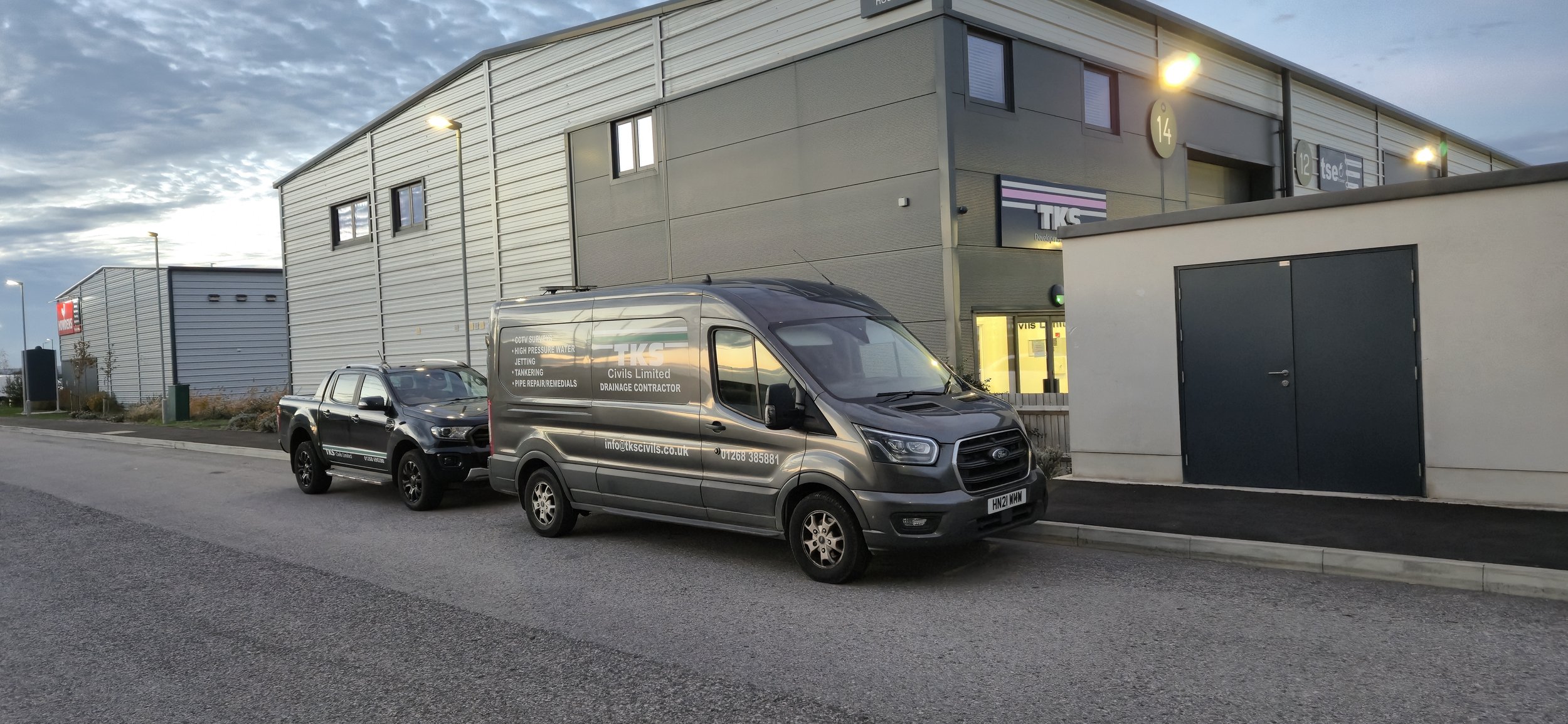 A street view outside a modern industrial building with parked service vehicles, including a grey van and a small black SUV, featuring company logos and contact information.