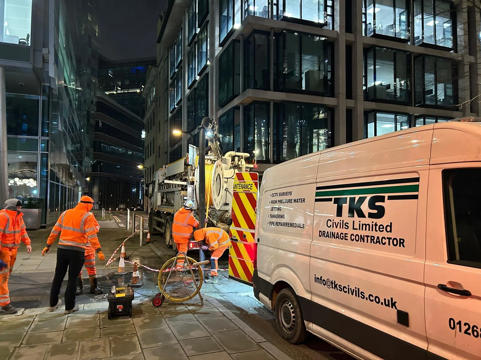 Construction workers in orange safety gear working on a maintenance project on a city street at night, with modern glass buildings in the background.