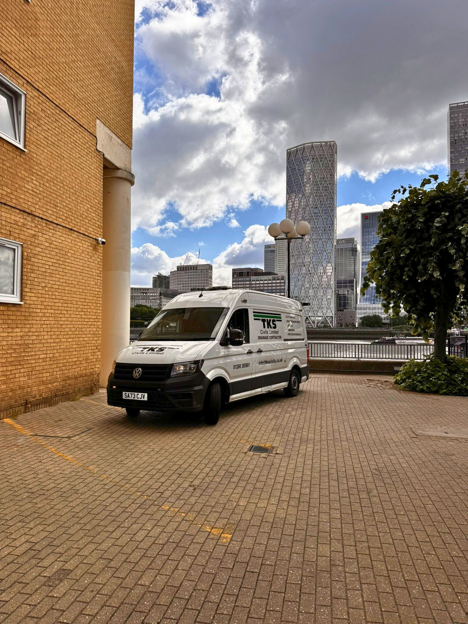 Cityscape with tall modern buildings, partly cloudy sky, parked white van in the foreground, brick building on the left, and a tree on the right.