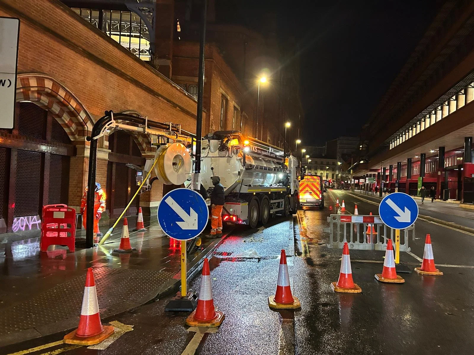 City street at night with road work and maintenance vehicles. Construction workers are present, with orange cones and barriers cordoning off the area. A large vacuum truck is visible, and workers are operating equipment. Streetlights illuminate the scene, and wet pavement reflects the lights.