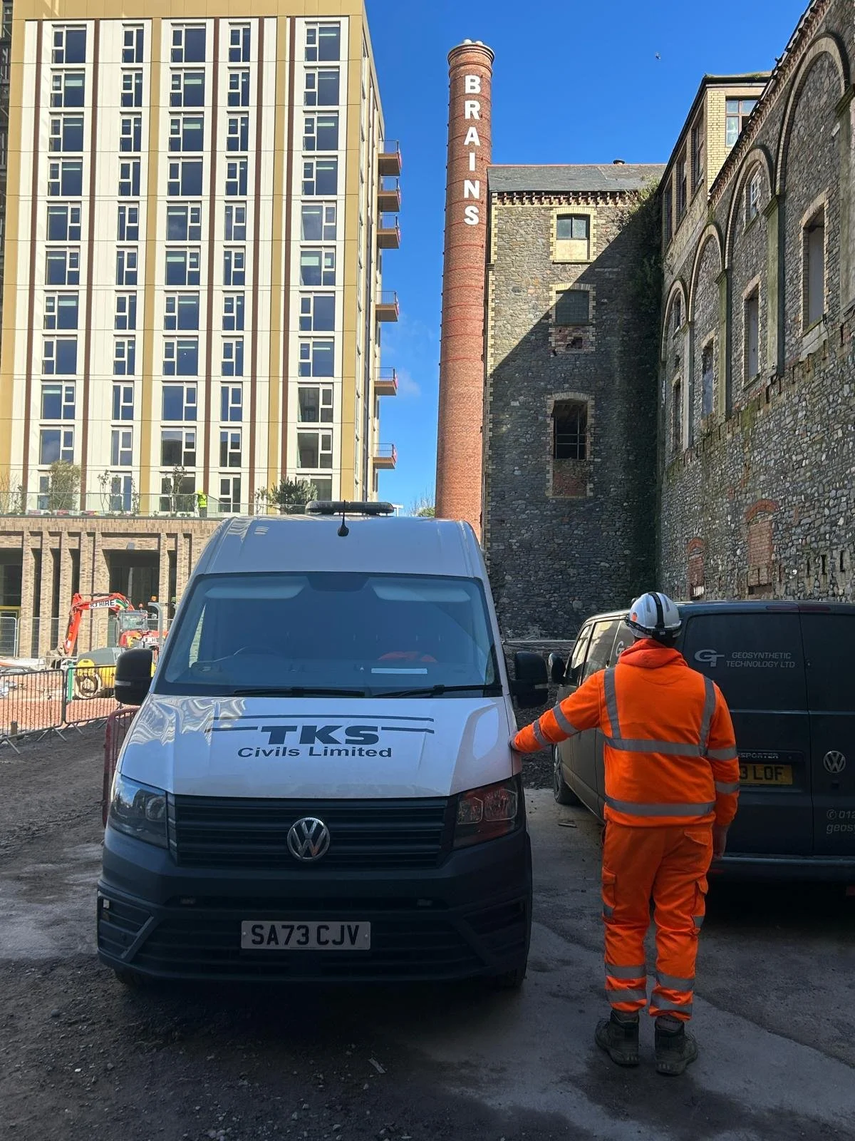 Construction worker in orange safety gear and helmet standing near construction vehicles on a site with a tall, modern building on the left and an older stone building with a tall brick chimney bearing the word 'BRAINS' on it in the background.