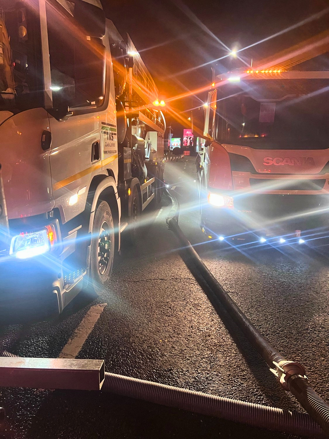 Nighttime scene of a fire truck and a bus on a wet road, with water hoses and emergency lights flashing.