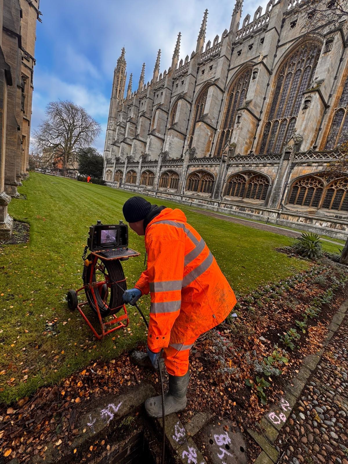A worker in an orange safety jacket kneels on the ground outside a historic cathedral, operating a surveying instrument connected to a laptop placed on a mobile stand. The cathedral has large stained glass windows and gothic architecture.